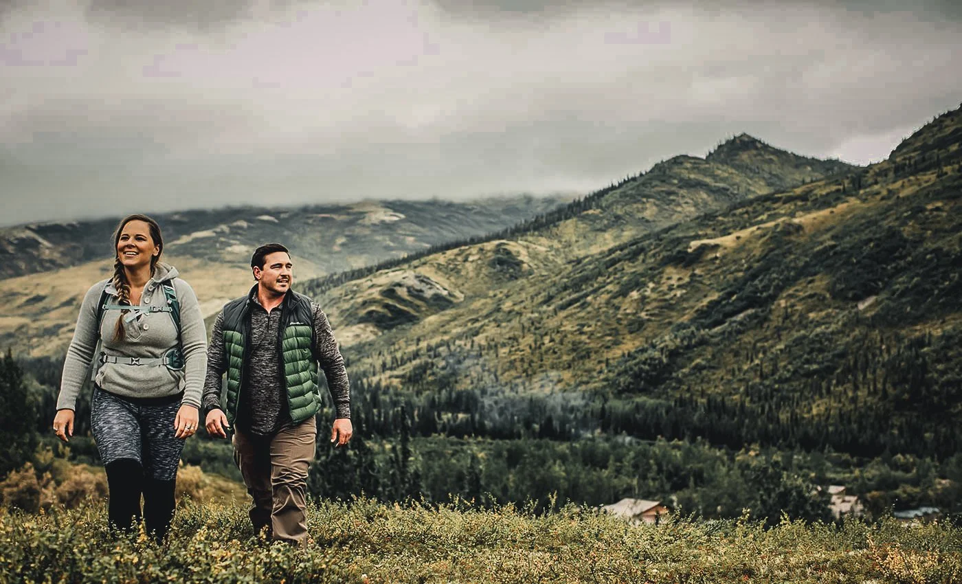 A woman and a man hiking together in a mountainous area, carrying backpacks, with a cloudy sky overhead.