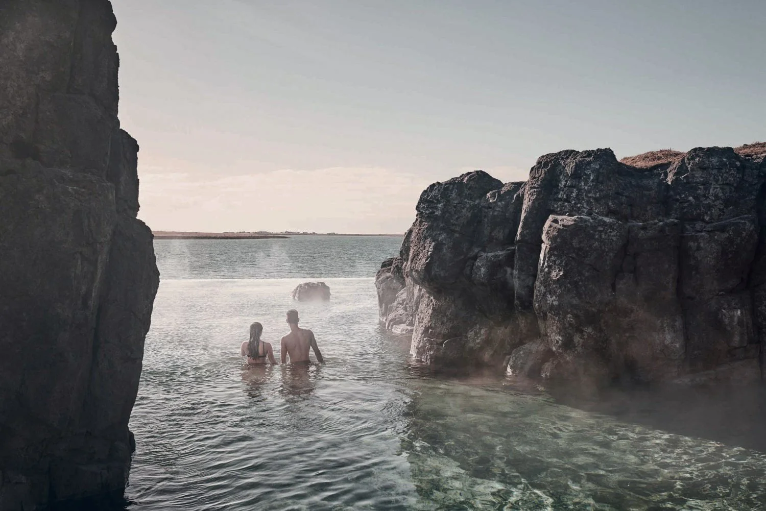 Two people, a man and a woman, standing in the water between large rocks with a body of water and horizon in the background