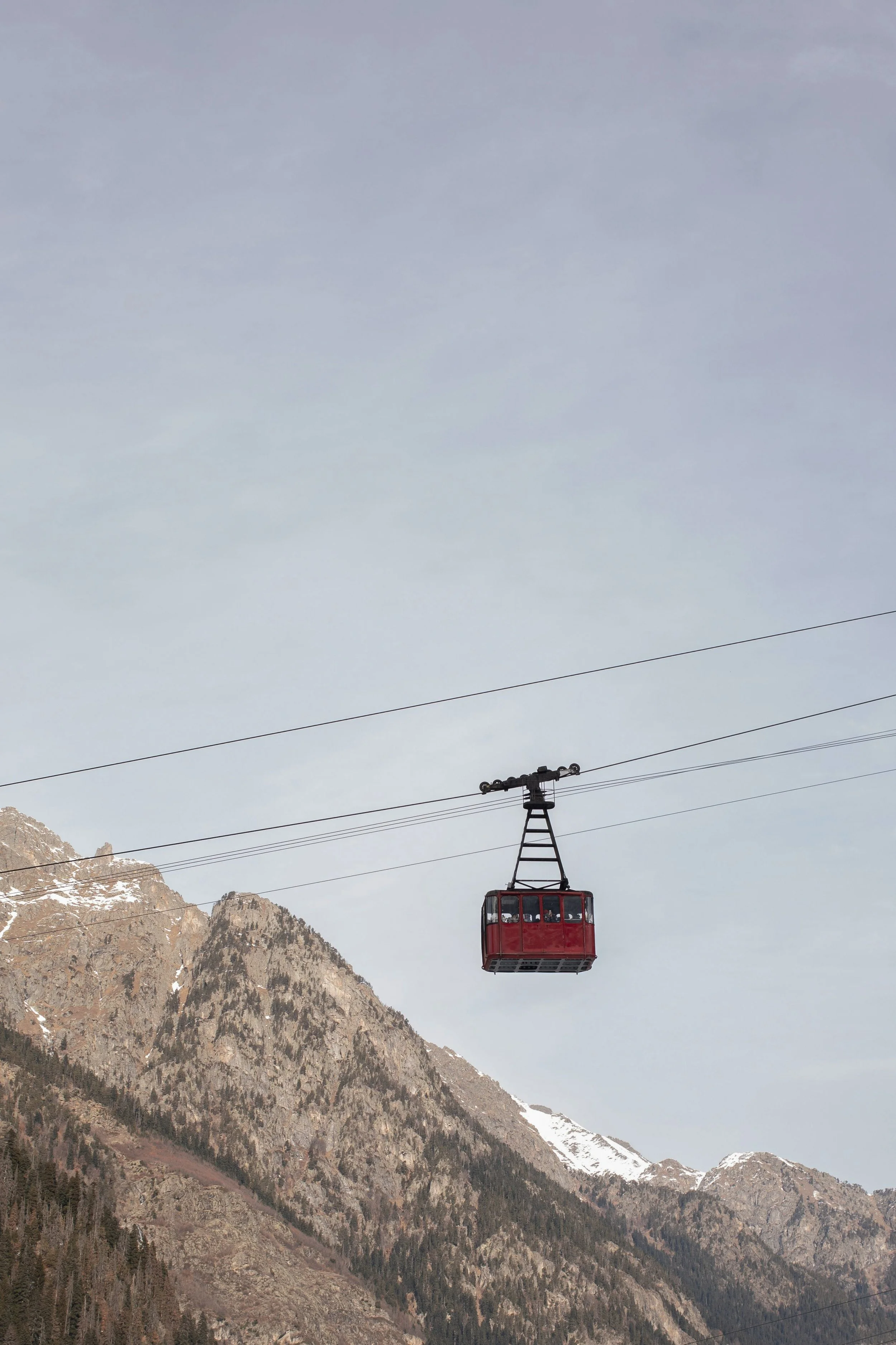 A red cable car traveling over a mountain landscape with snow-capped peaks in the background.
