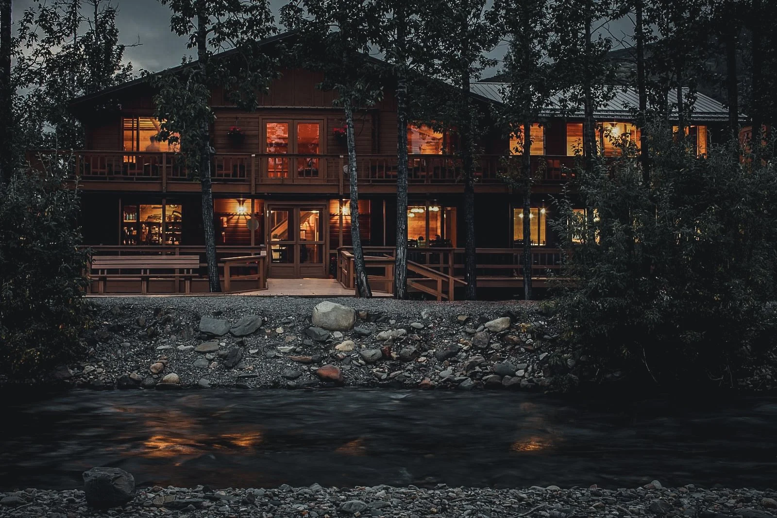 A multi-story house with large windows, wooden exterior, and balconies, situated behind trees along a rocky riverbank at dusk or night with warm interior lighting visible through the windows.