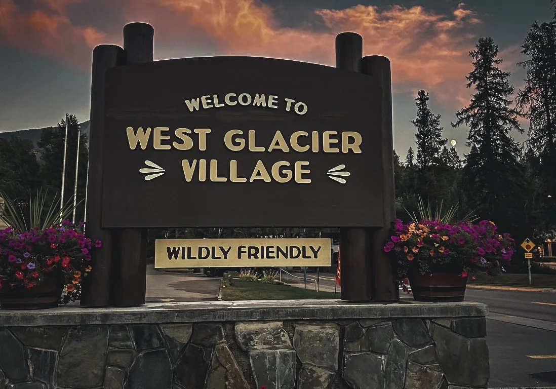 Sign welcoming visitors to West Glacier Village, describing it as wild and friendly, with flowers on either side and a mountain landscape in the background.