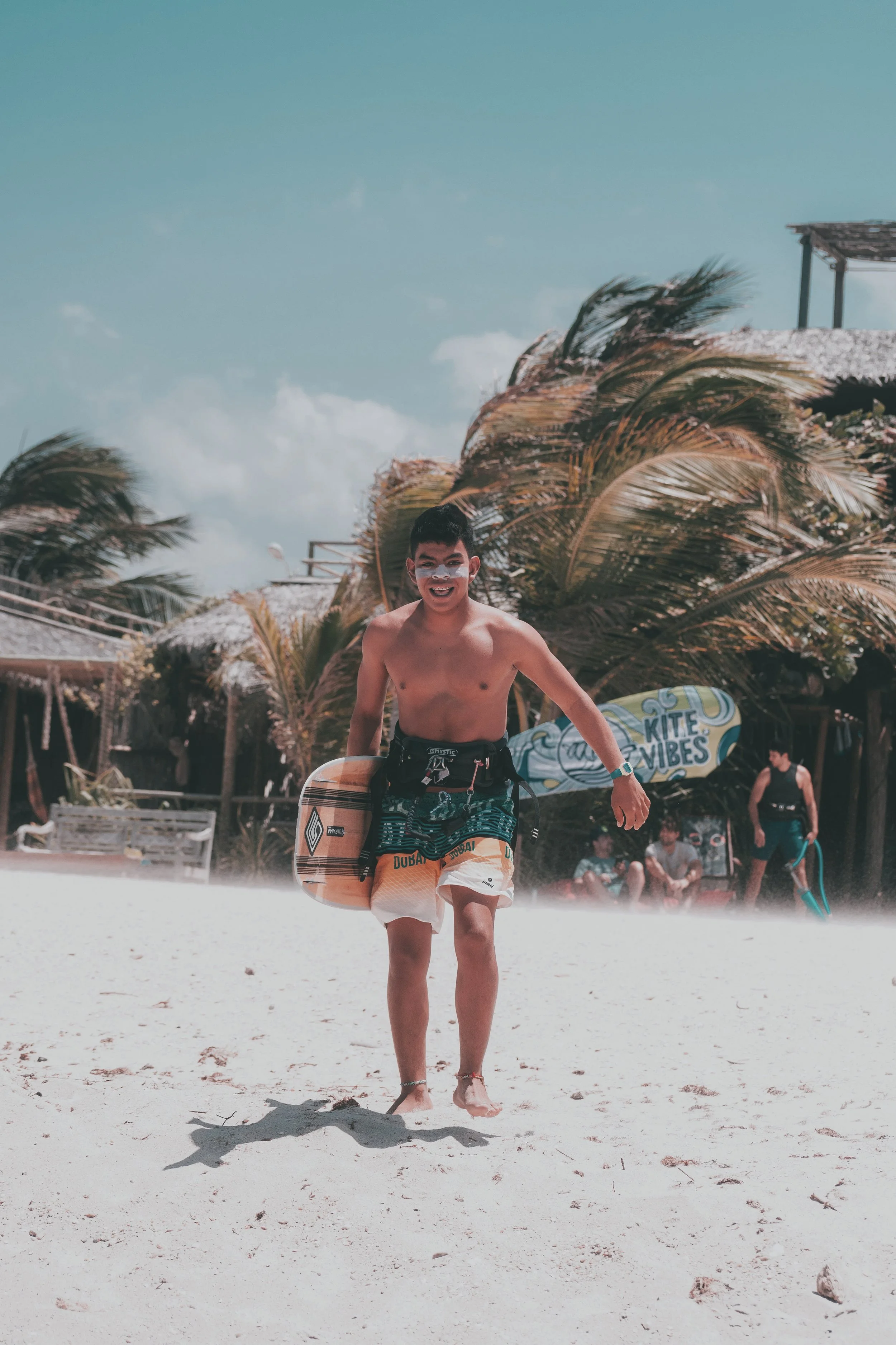 A young man smiling and running on a sandy beach, carrying a surfboard, with palm trees and beachside structures in the background under a partly cloudy sky.