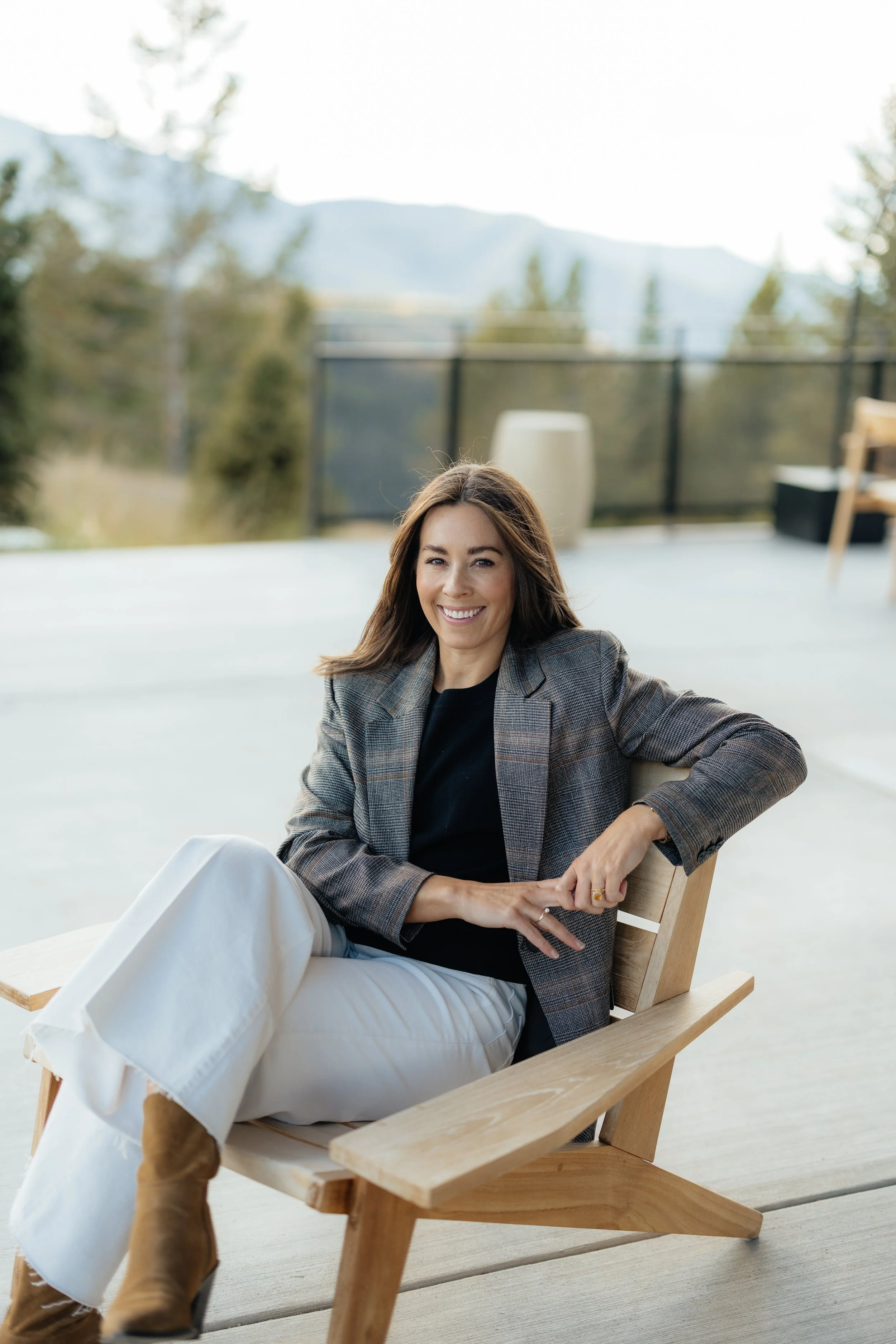 A woman with long brown hair smiling, sitting on a wooden chair outdoors with mountains and trees in the background.