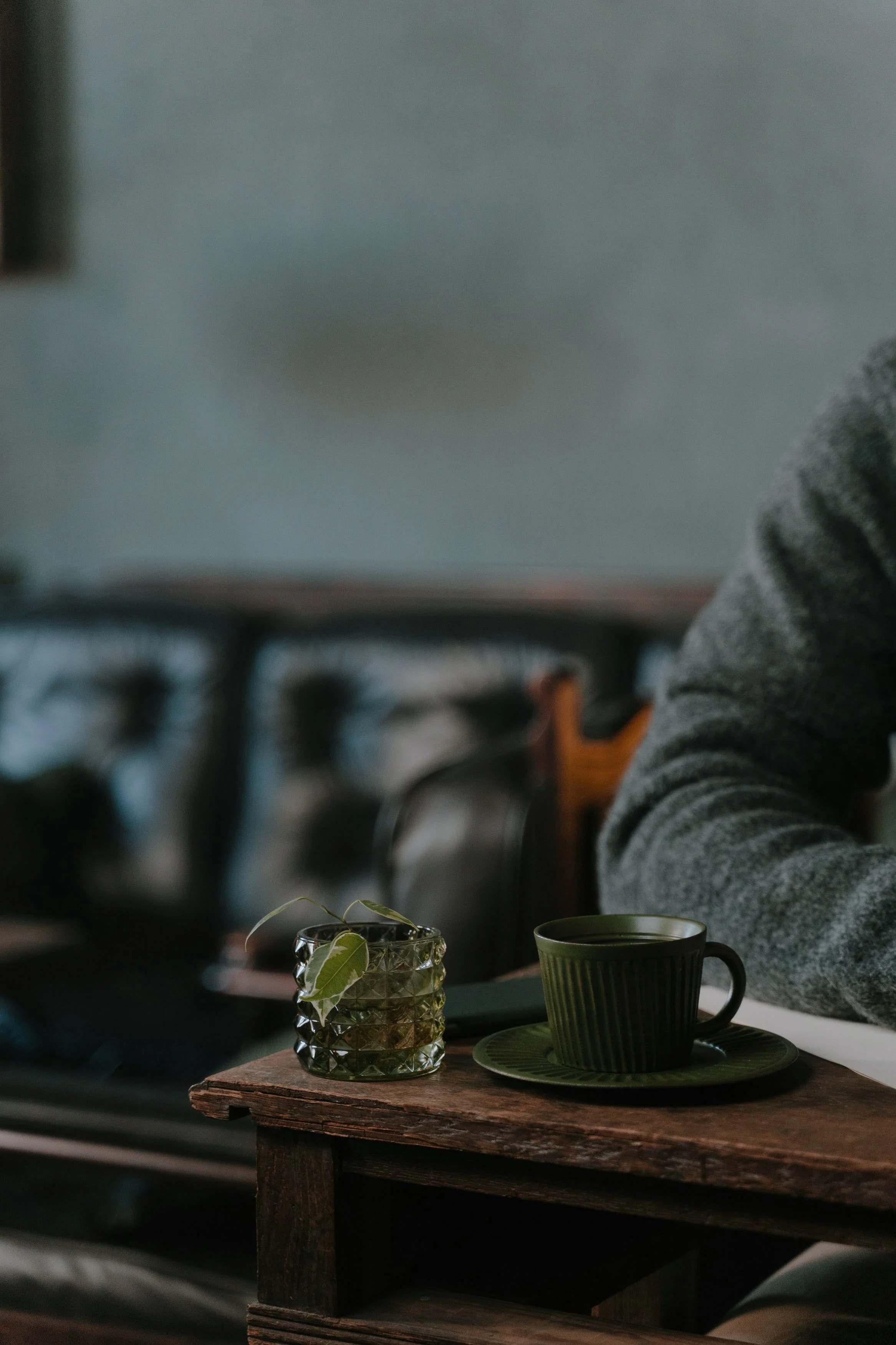 A wooden table with a dark green cup on a matching saucer and a glass container with leaves inside, set against a blurred background with a person wearing a gray sweater.
