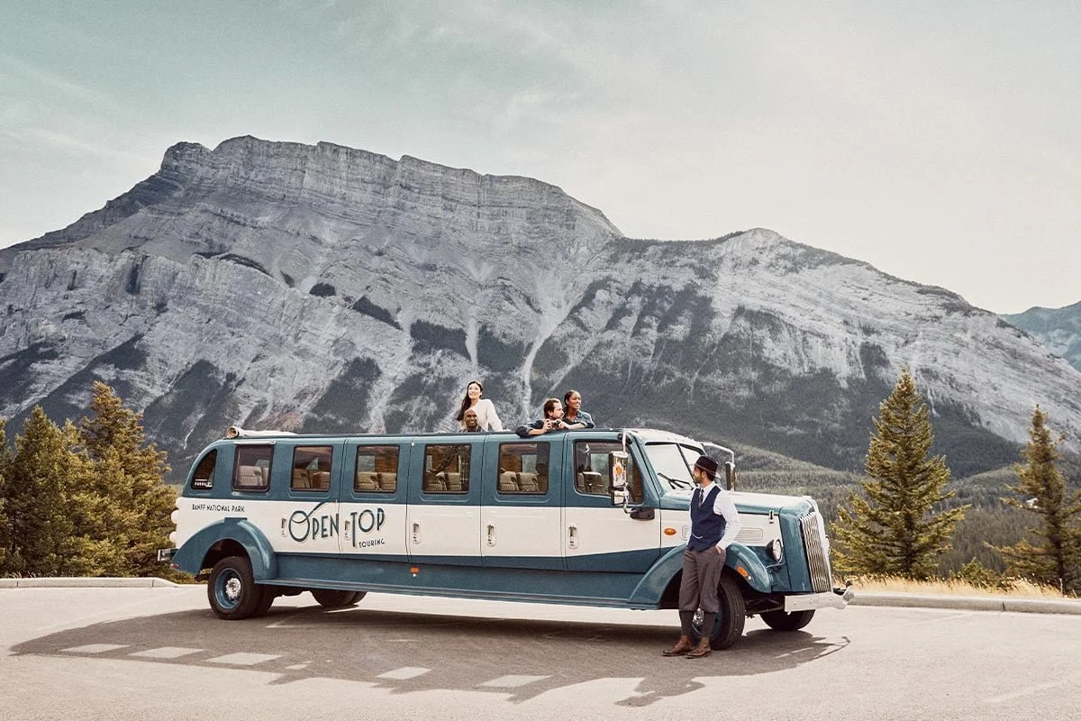 Tourists on a vintage blue and white bus in a mountainous landscape with trees, and a tour guide standing outside, wearing a vest and hat.
