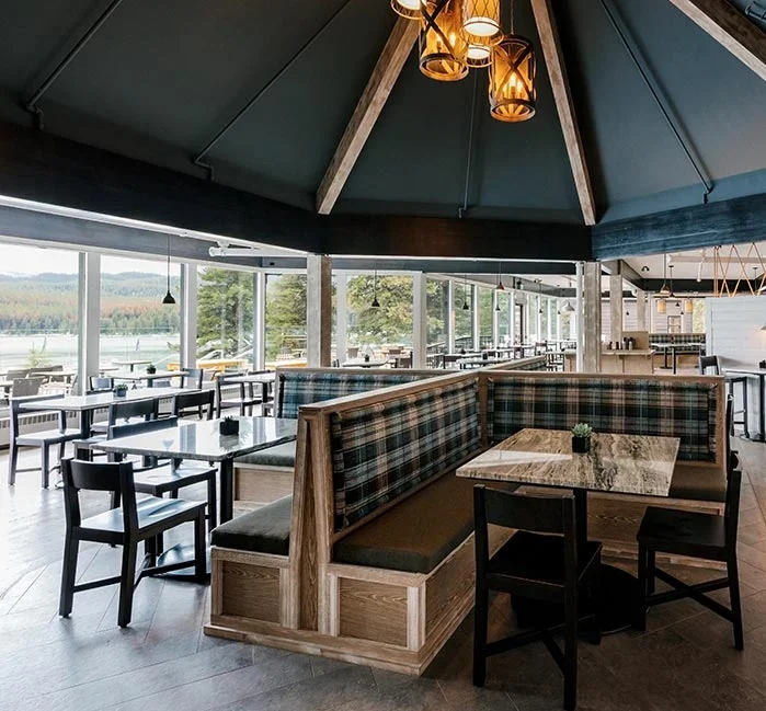 Interior of a restaurant with large windows, wooden tables and chairs, and a green ceiling with hanging lights.