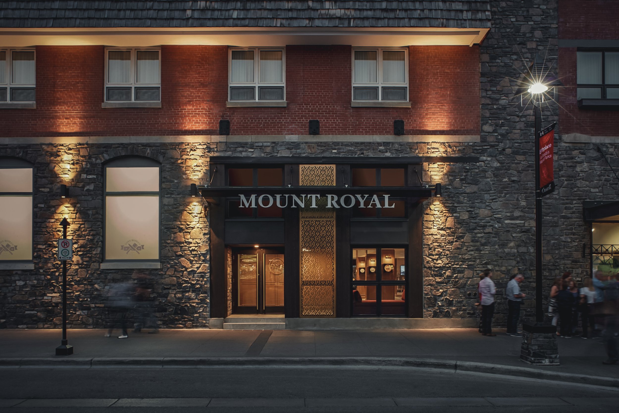 The entrance of Mount Royal hotel illuminated at night with a stone and brick facade, several people standing outside, and a streetlamp nearby.