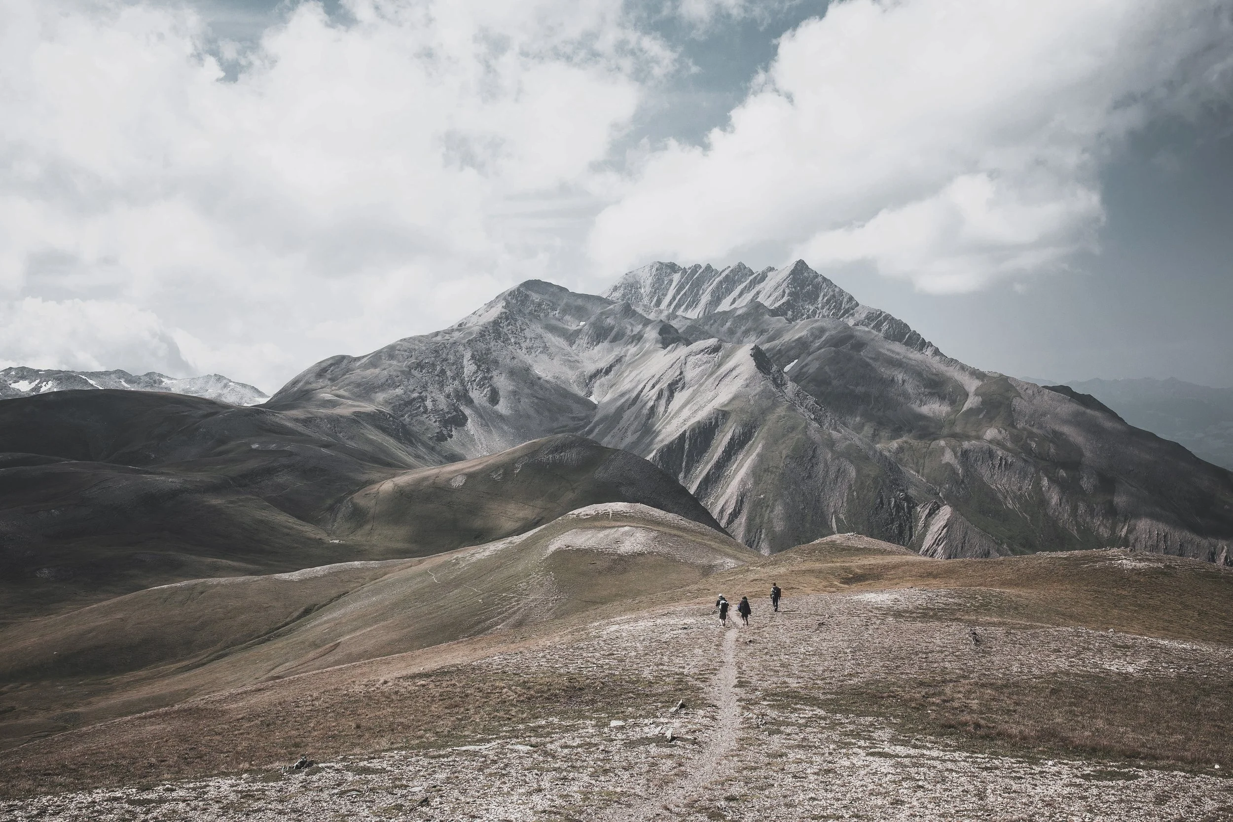 Three hikers walking on a mountain trail with rolling hills and rugged snow-capped mountains in the background under a cloudy sky.