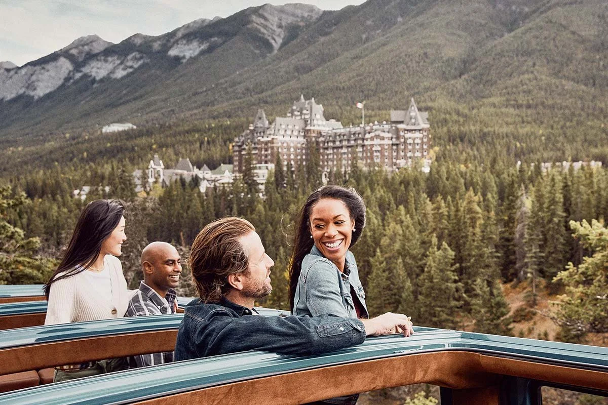 A group of four people enjoying a ride on a scenic train with a backdrop of mountains, trees, and a castle in the distance.