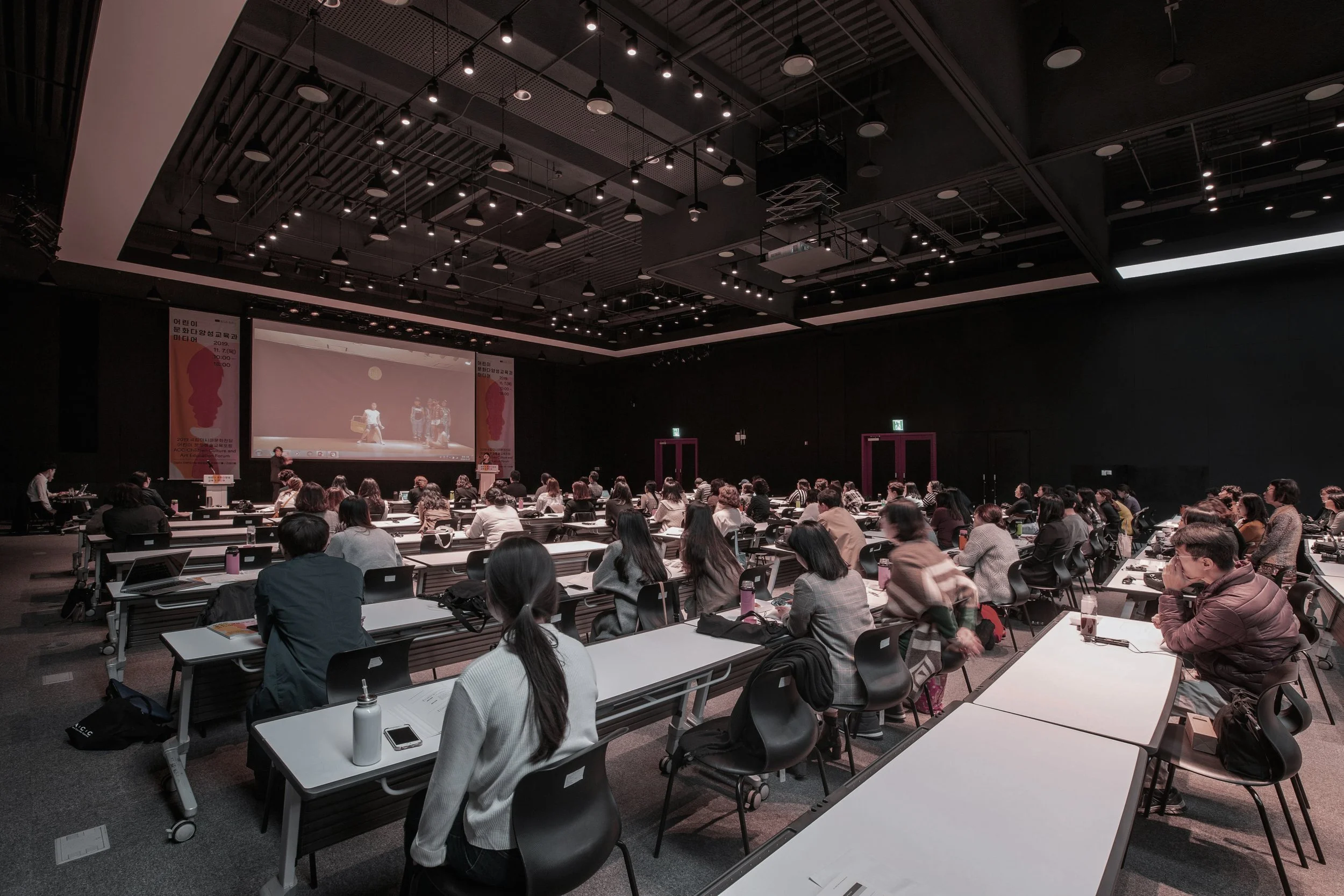 A large conference room filled with people seated at tables, watching a presentation on a big screen at the front of the room. The room has a dark ceiling with numerous lights.