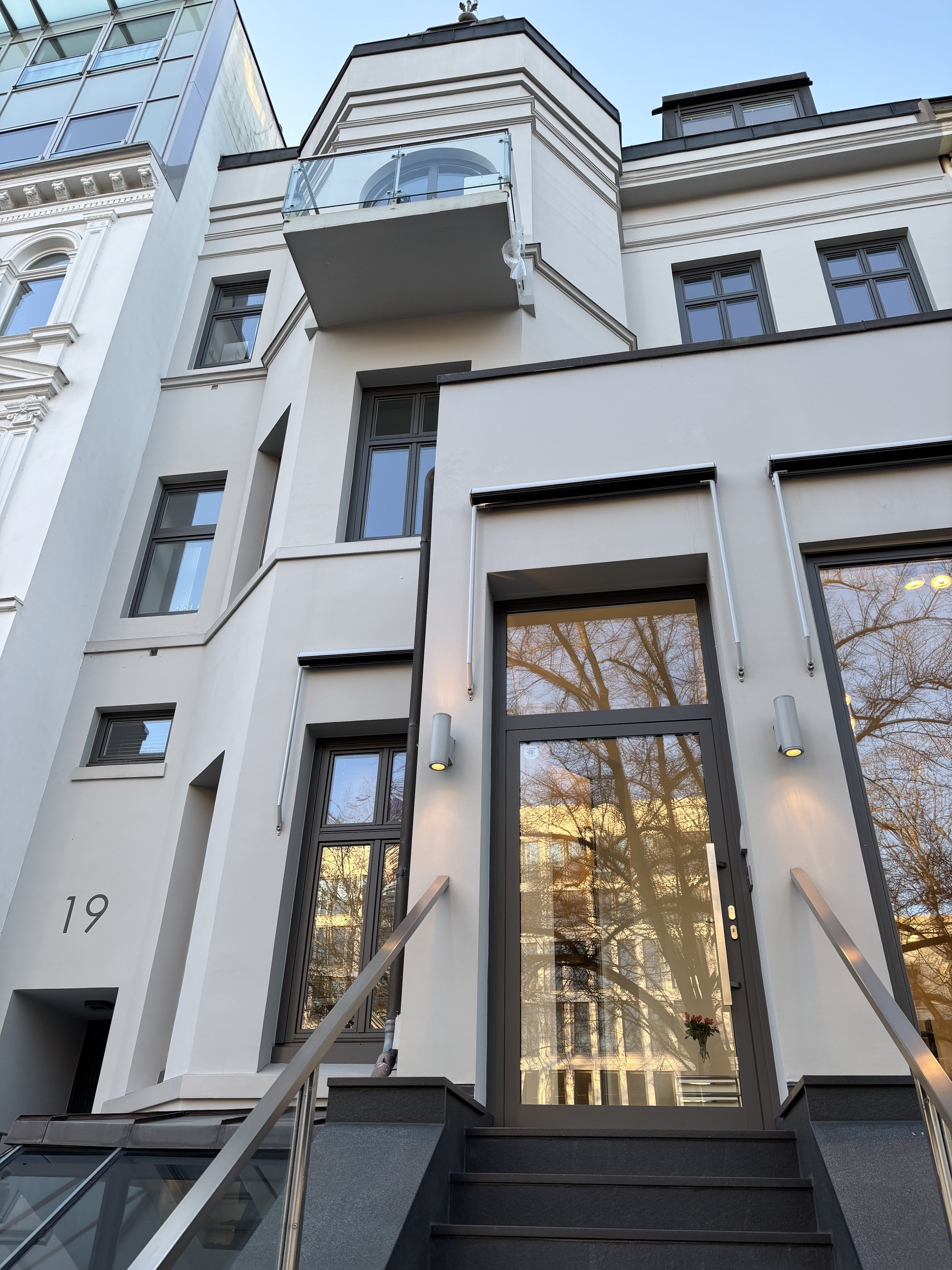 Altbauobjekt in grau sichtbar. Zudem ein blauer Himmel. Es ist Licht durch die Fenster zu sehen. Die Fenster sind anthrazit. Das Gebäude selbst hellgrau. Es hat sehr viele Versprenge, aber gradlinig und schick.