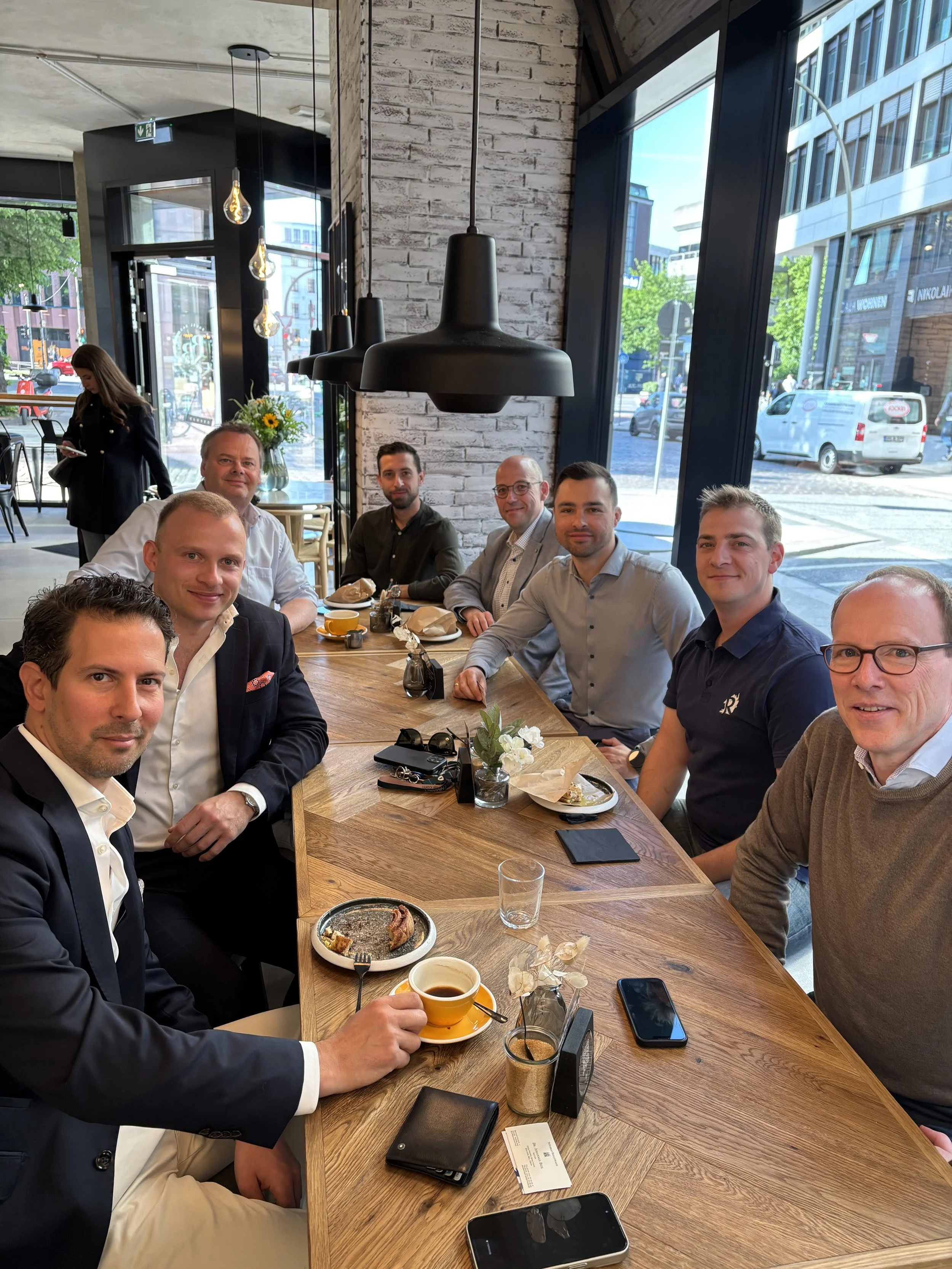 Eine Gruppe von neun Männern sitzt um einen großen Holztisch in einem modern eingerichteten Café, einige trinken Kaffee, andere haben Essen vor sich.