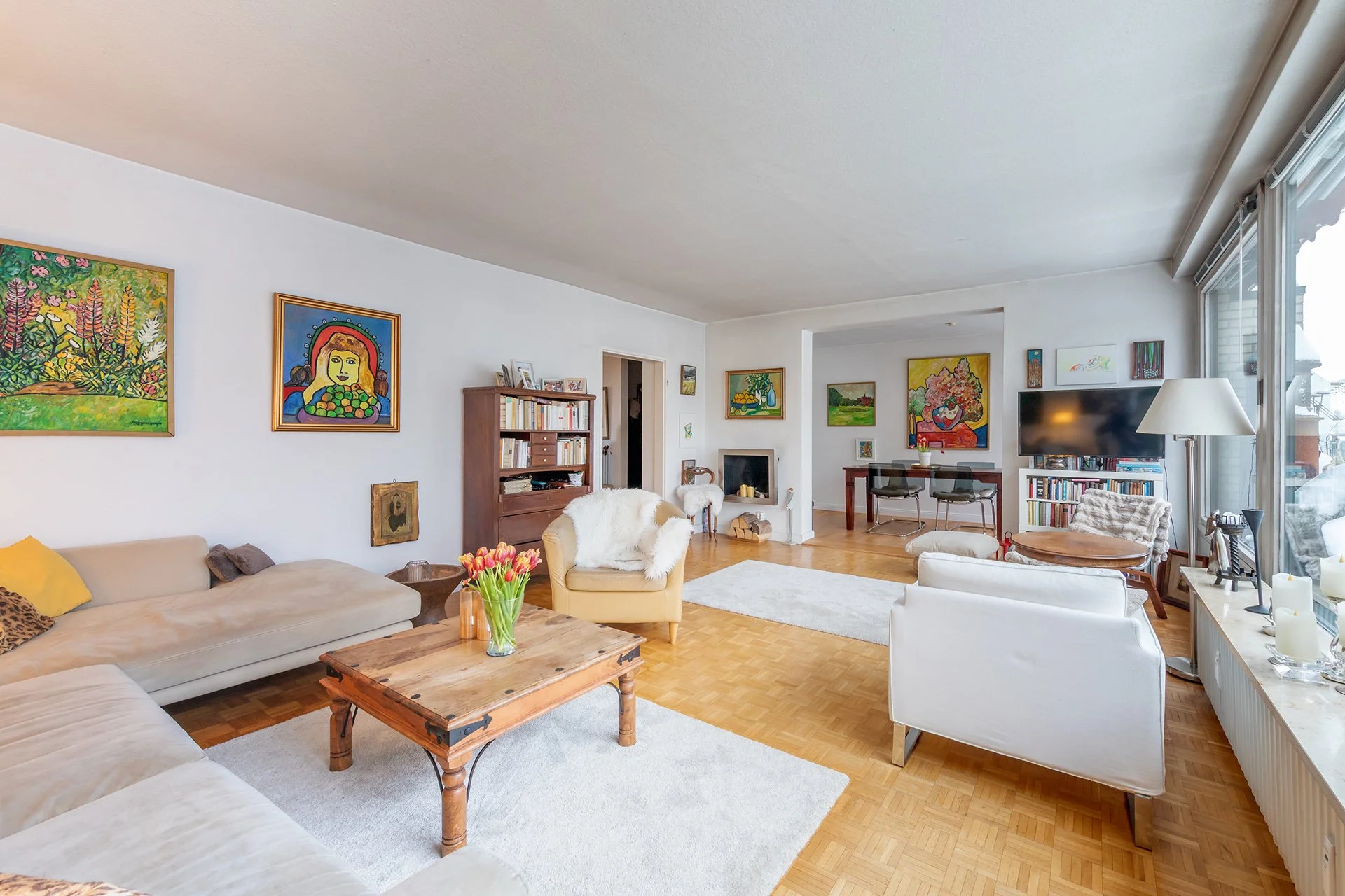 Wohnzimmer mit Parkett, Sesseln, Sofa, Tischen und bunten Wildern. Überall weiße Wände und rechts ein Fenster