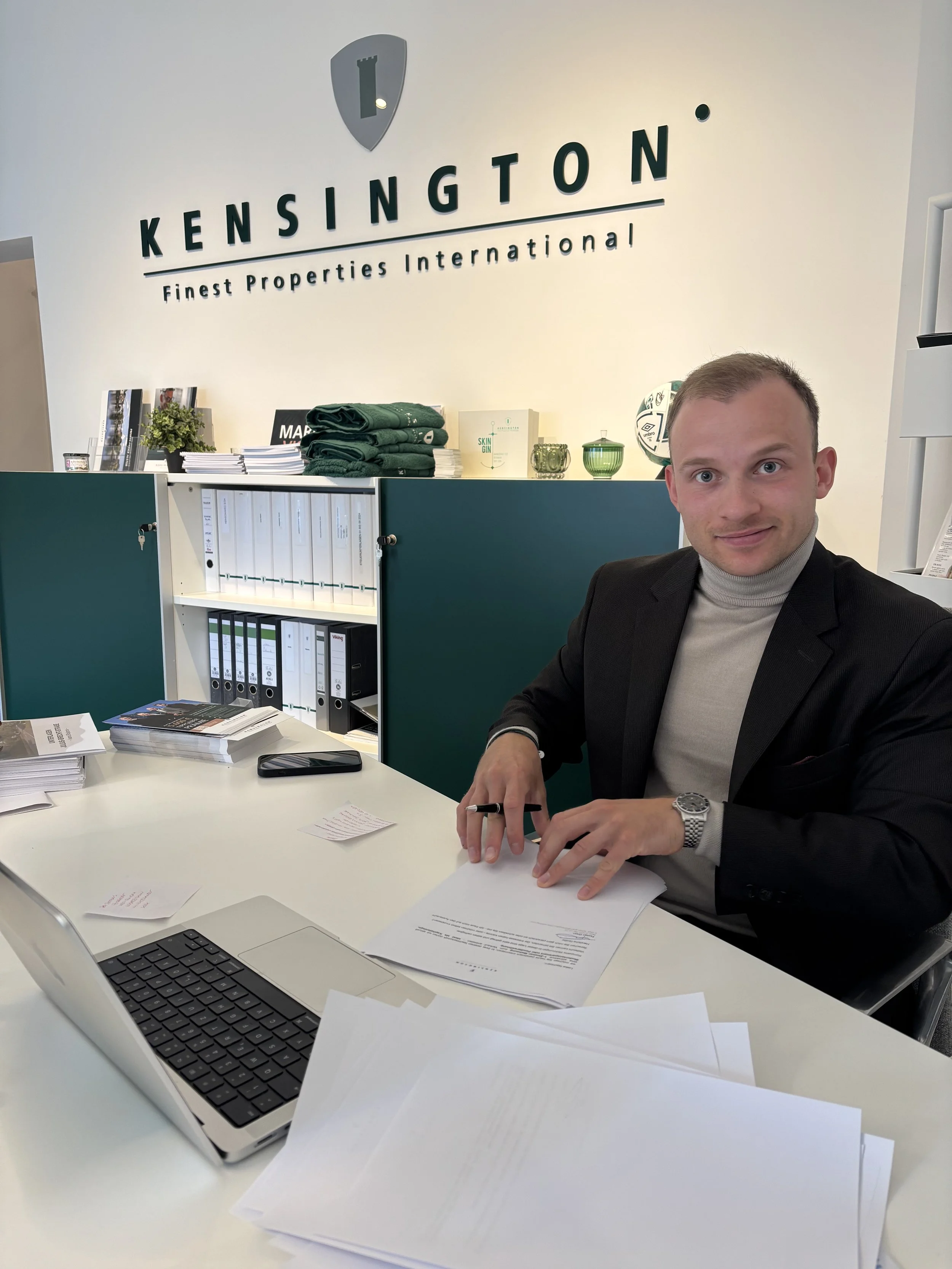 Ein Mann in einem schwarzen Anzug sitzt an einem weißen Schreibtisch, auf dem sich ein Laptop, Papier, Notizzettel und ein Smartphone befinden. Im Hintergrund die Firmenbeschriftung 'KENSINGTON Finest Properties International' an der Wand.