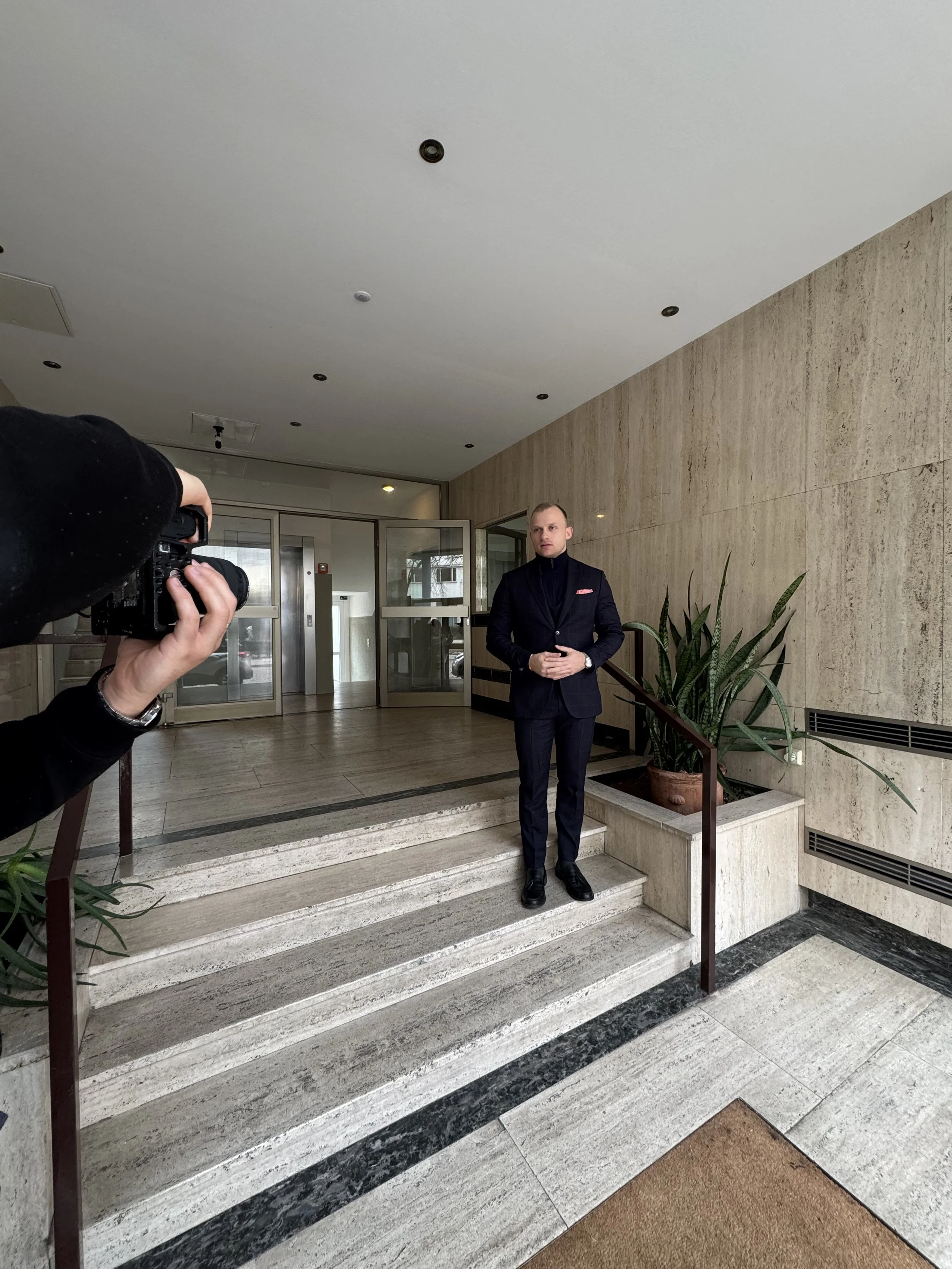 Ein Mann in dunkler Anzug steht auf den Treppen vor einem Gebäude, während eine andere Person ein Foto mit einer Kamera macht.