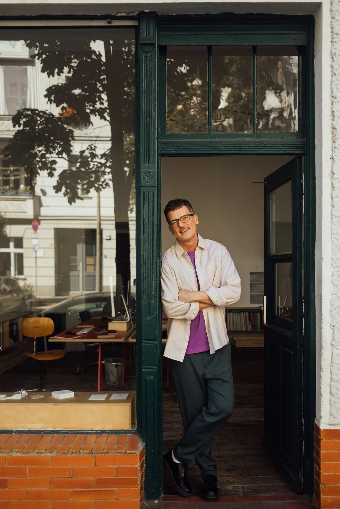 A man with glasses, wearing a white shirt over a purple t-shirt, stands in an open doorway with his arms crossed, smiling. Inside the room behind him, there are books, a desk, and chairs. The outside scene reflects trees and buildings.