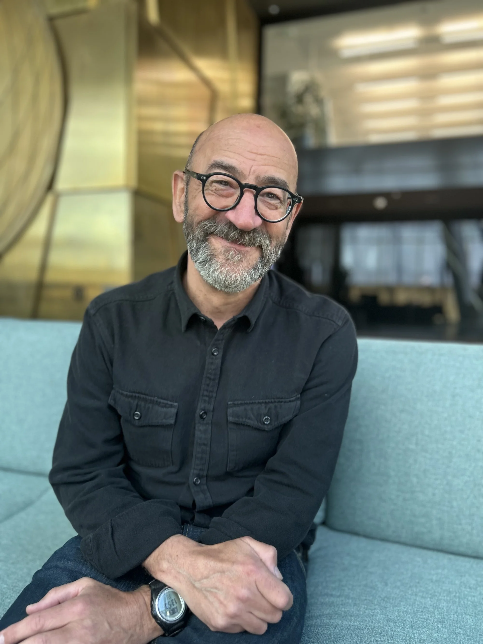 Lächelnder Mann mit Brille und grauem Bart sitzt auf einem hellblauen Sofa in einem modernen Raum mit Holzdekor und Glasbalustrade.