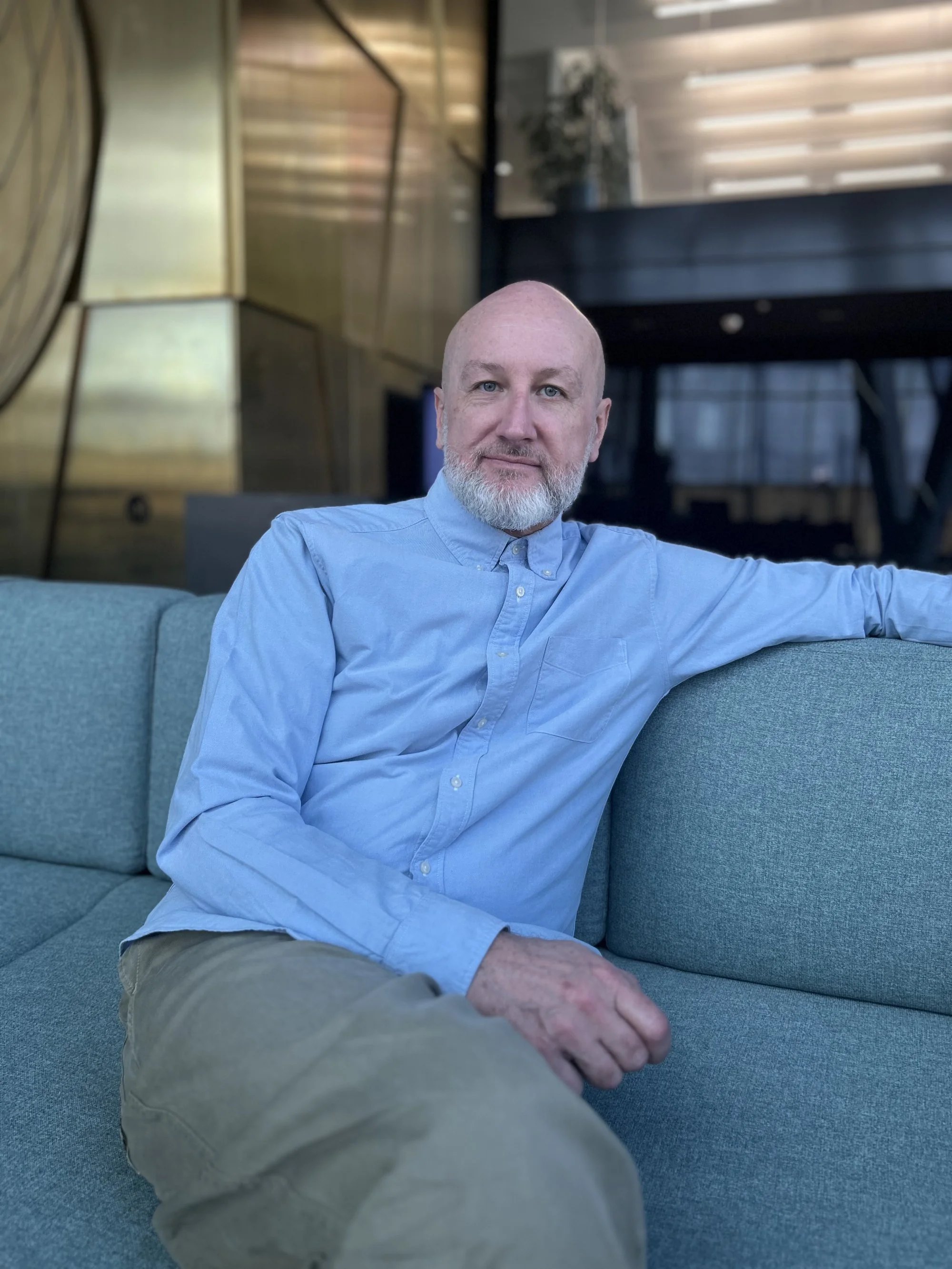 Ein älterer Mann mit Glatze und grauem Bart sitzt entspannt auf einem blauen Sofa in einer modernen, stilvollen Umgebung.