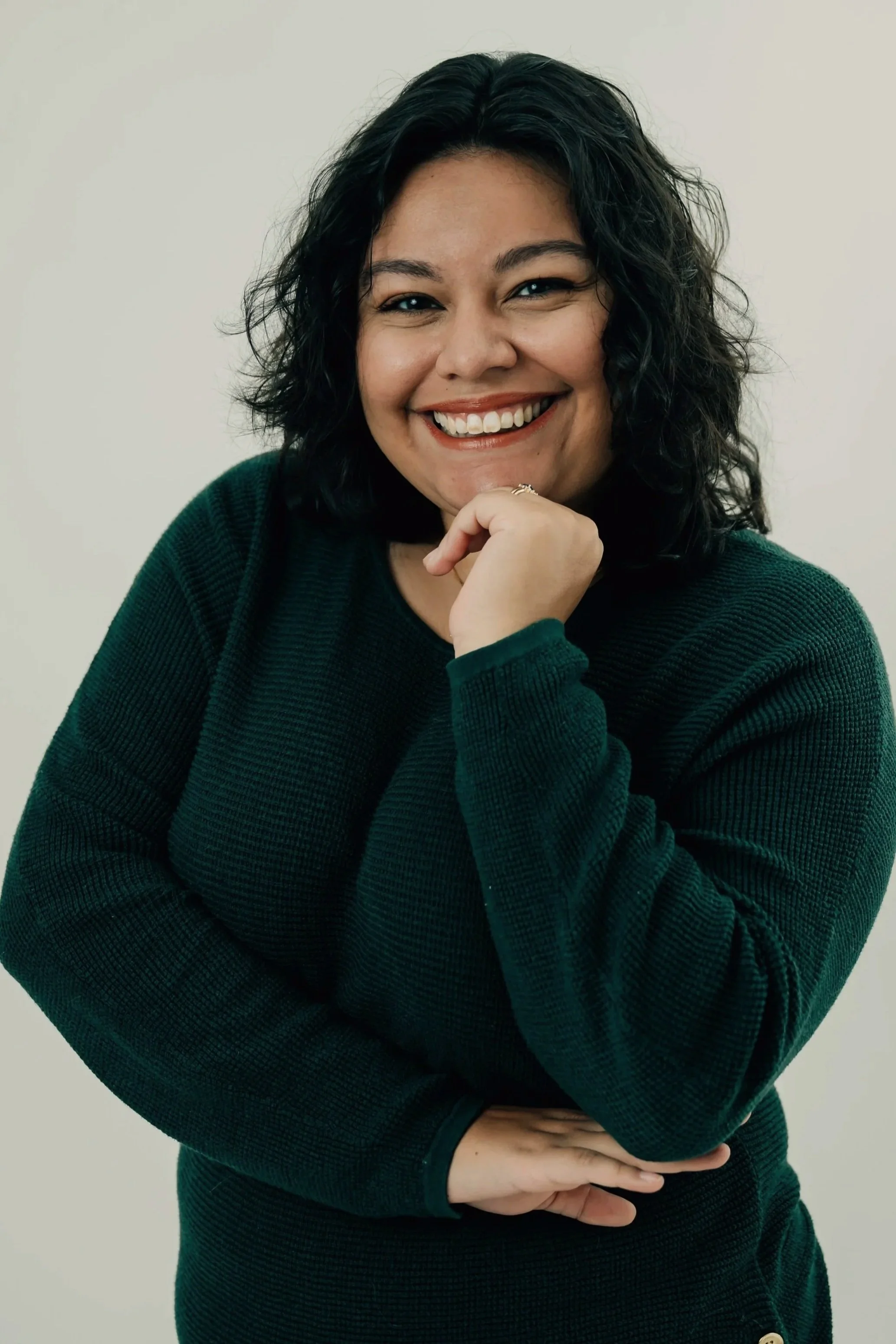 Gabby Camacho, virtual assistant, with curly black hair, smiling, wearing a dark green sweater, against a plain light background.