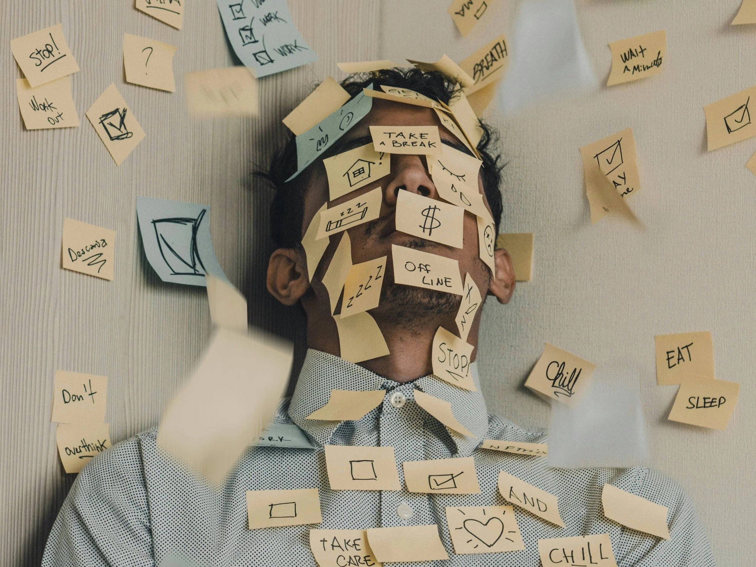 man up against the corner of a wall, with sticky notes on his face, body, and walls. Sticky notes have images, checklists, and money to express stress and burnout