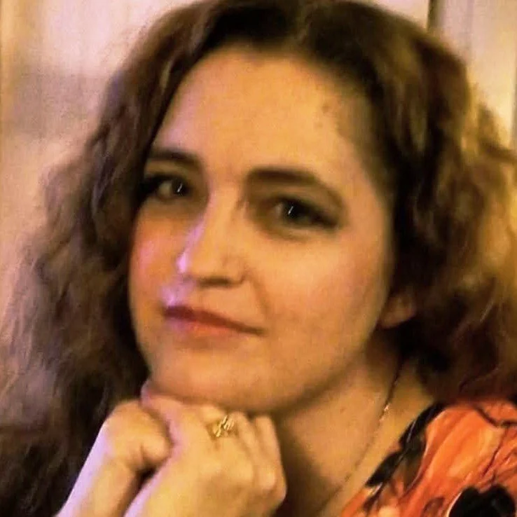 A woman with wavy brown hair posing with her chin resting on her hand, looking at the camera with a slight smile, wearing orange and black clothing.