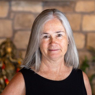 A woman with shoulder-length gray hair wearing a black sleeveless top, standing in front of a brick wall with some decorative plants.
