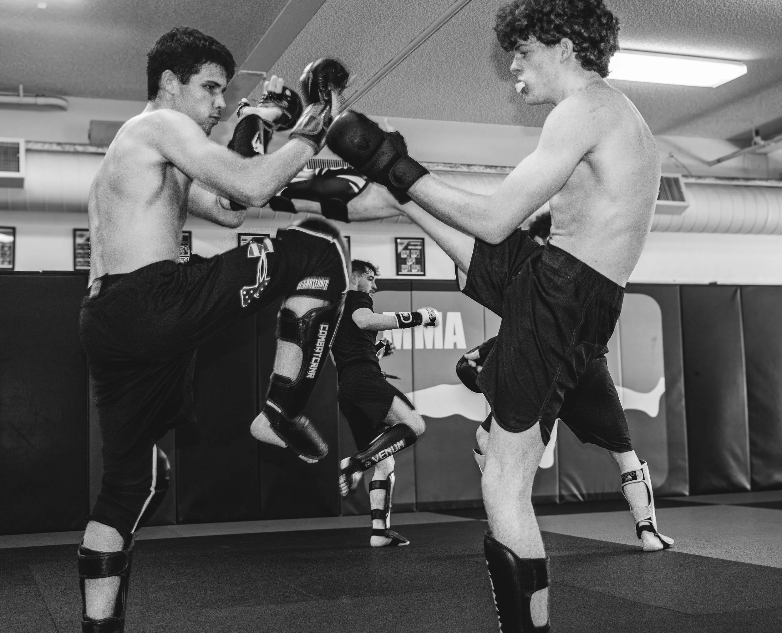 Two young males practicing mixed martial arts, with one kicking and the other blocking, in a gym with padded walls and two other fighters training in the background.