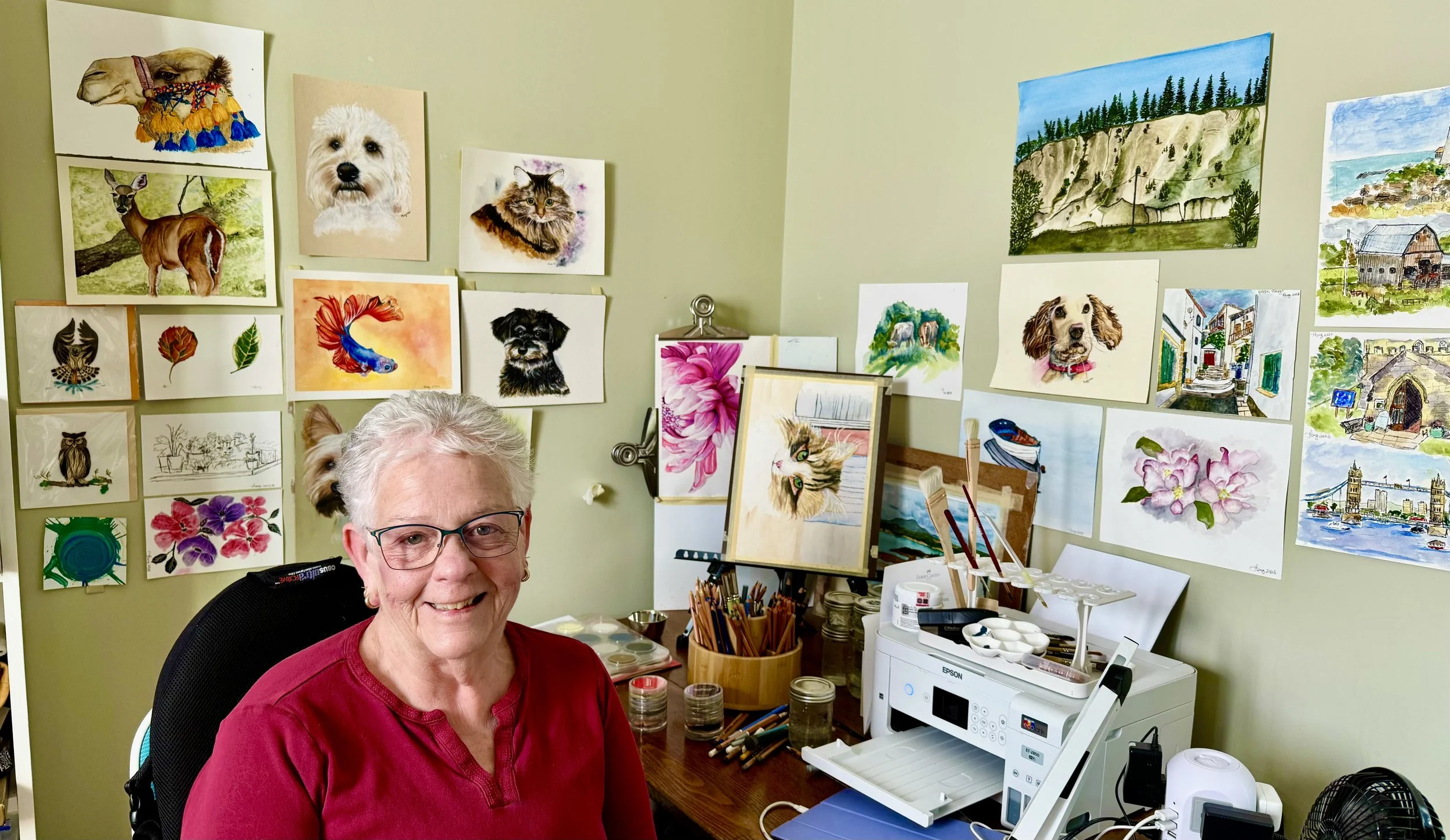 An elderly woman with gray hair and glasses smiling in an art studio filled with colorful paintings of animals, landscapes, and flowers on the walls.