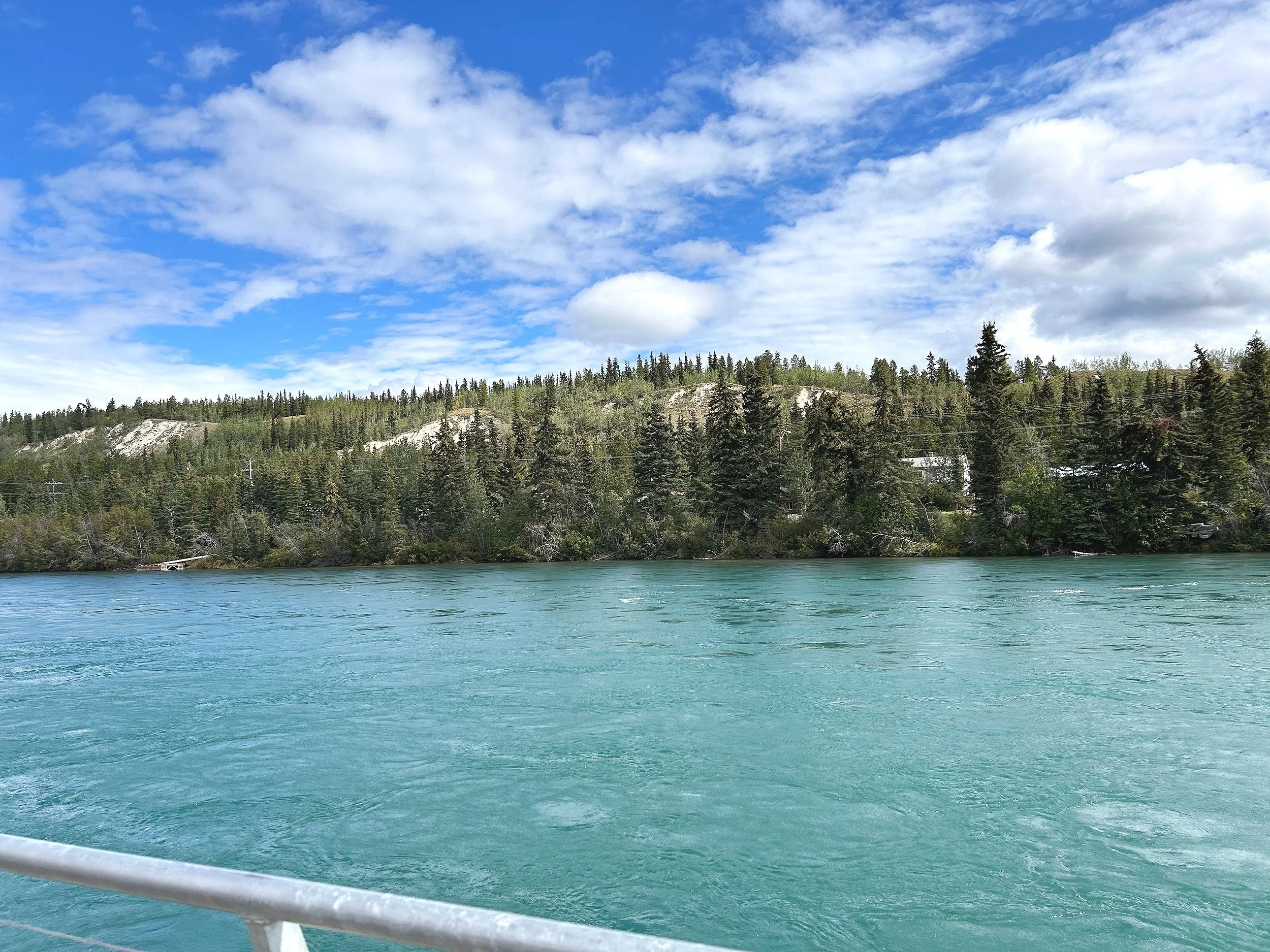 A body of turquoise water with a forested hillside in the background under a partly cloudy blue sky.