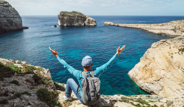 Person sitting on rocky cliff overlooking the ocean with arms raised, surrounded by rugged cliffs and clear blue water.