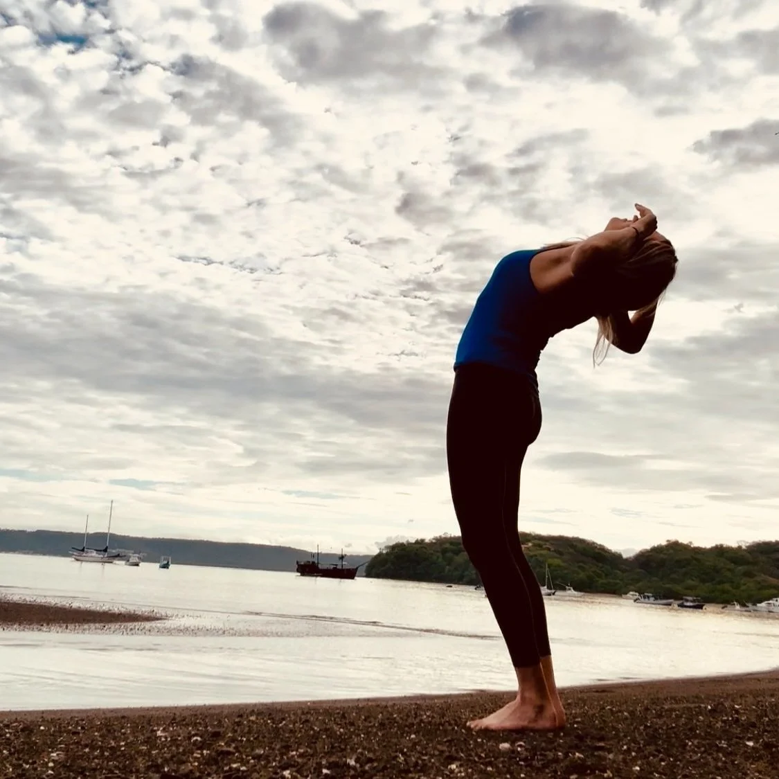 A person practicing yoga on a beach at sunset or sunrise, with water and boats in the background, standing in a forward bend pose with hands behind their head.