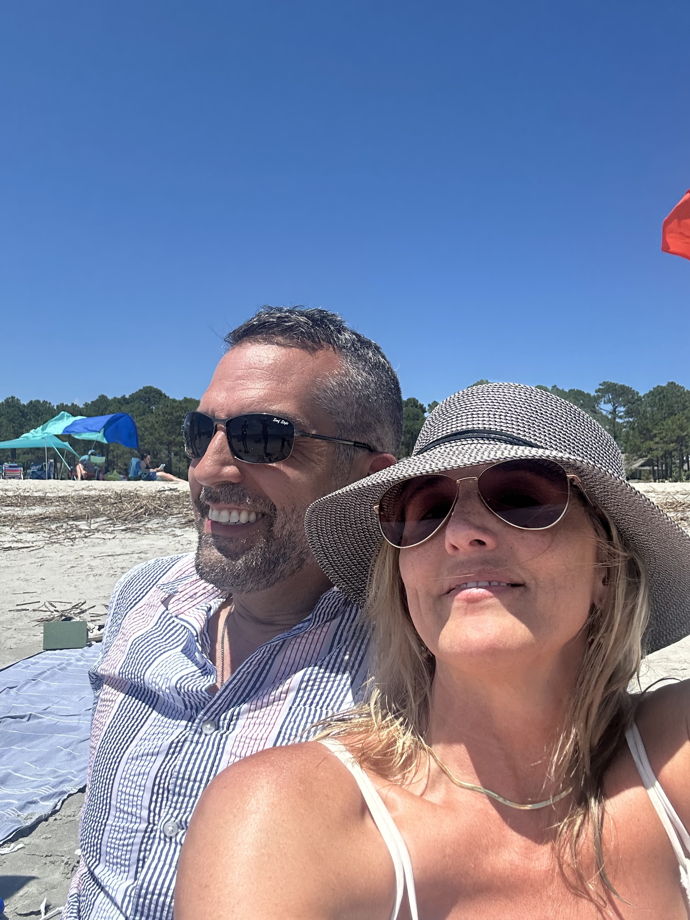 A man with sunglasses and a smiling woman wearing a sunhat and sunglasses, at the beach on a sunny day.