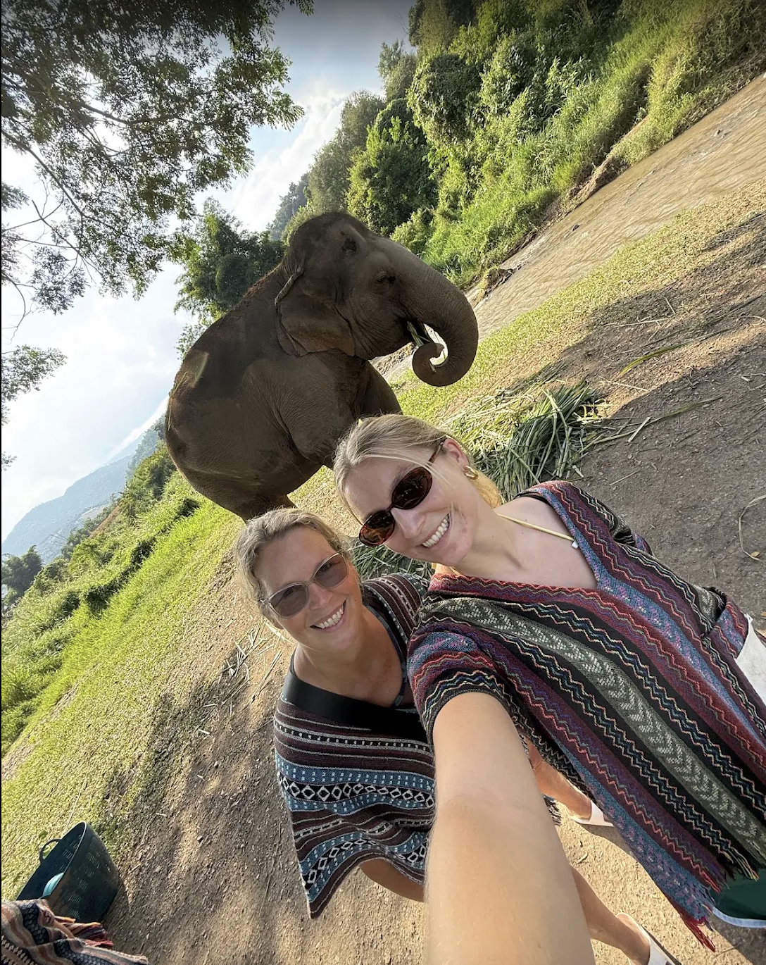 Two women taking a selfie with an elephant in a green, hilly outdoor setting, likely a jungle or nature reserve.