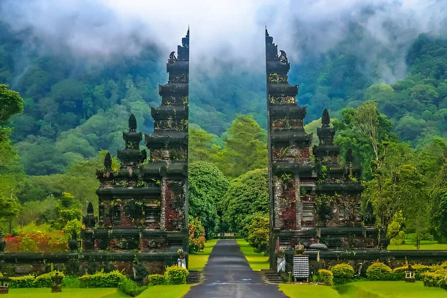 Stone entrance gates surrounded by lush green gardens and trees, with misty mountains in the background.