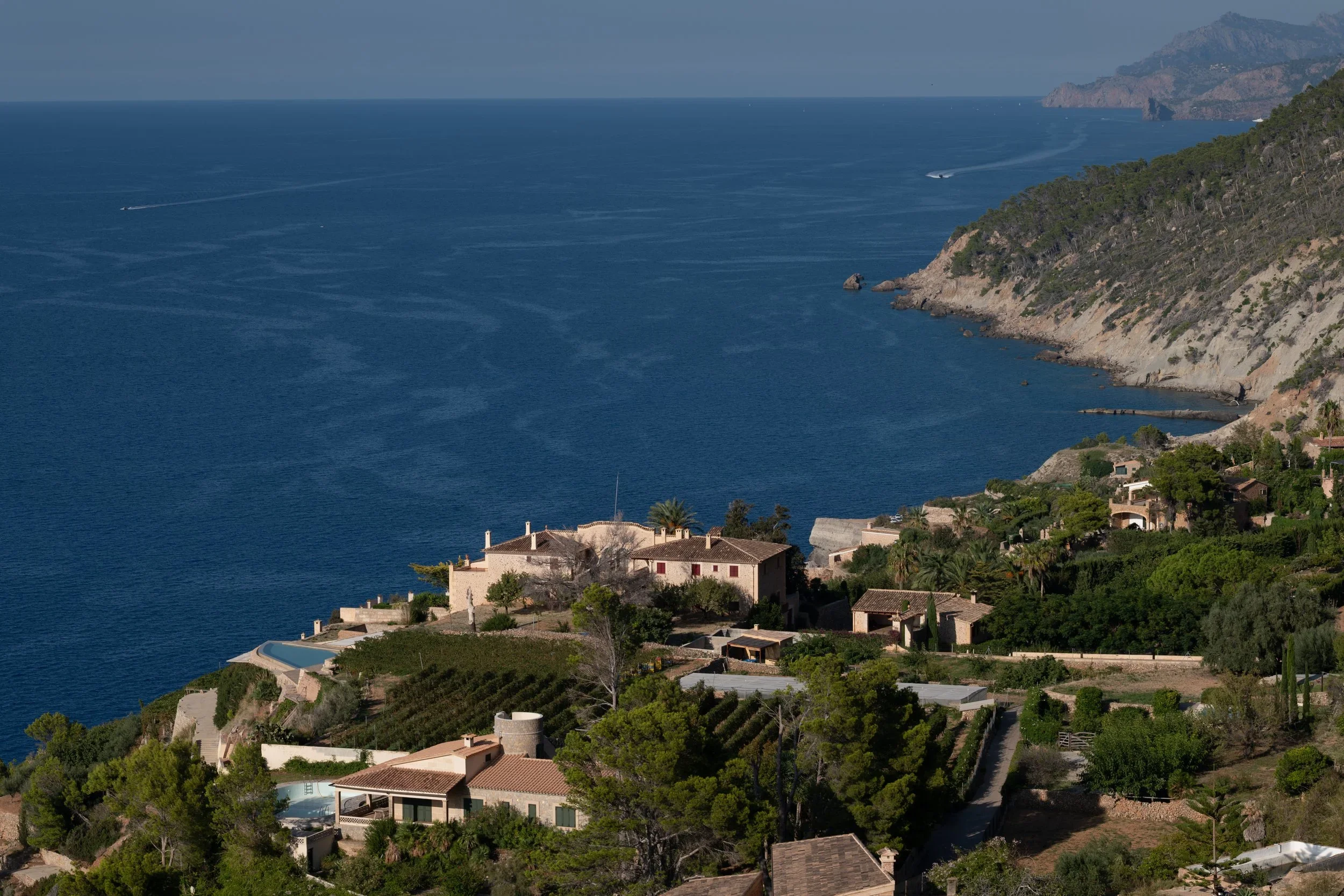 Coastal residential area with houses, trees, and a hillside overlooking the ocean, with boats visible on the water and distant mountains in the background.