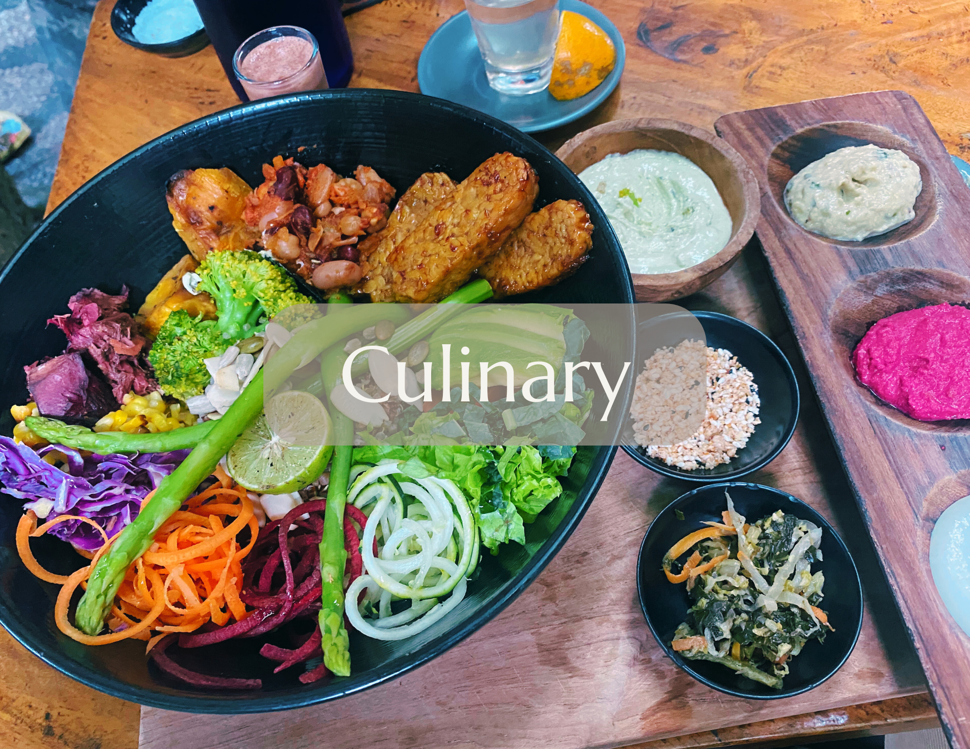 A colorful platter of vegan dishes including various vegetables, hummus, guacamole, and plant-based meats on a wooden table with small bowls of sauces and sides.