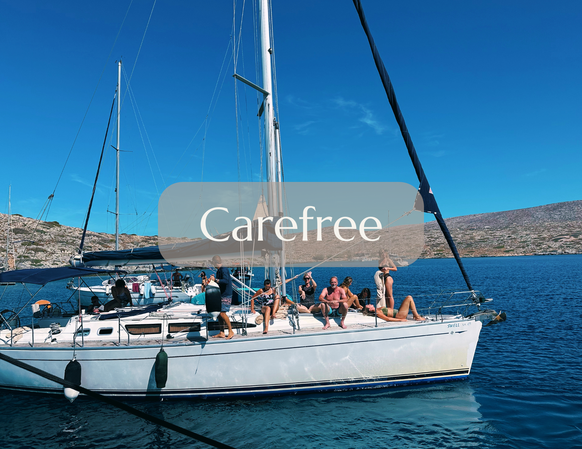 Group of people relaxing and enjoying the sun on a sailboat anchored in a calm body of water, with rocky hills in the background and a clear blue sky.