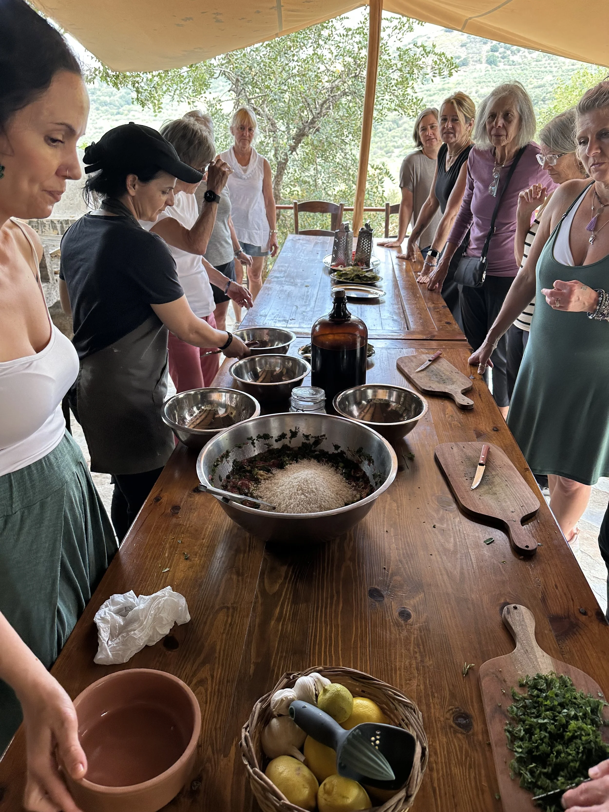 A group of women gathered around a wooden table outdoors, preparing ingredients with bowls of herbs, lemons, garlic, and kitchen tools.
