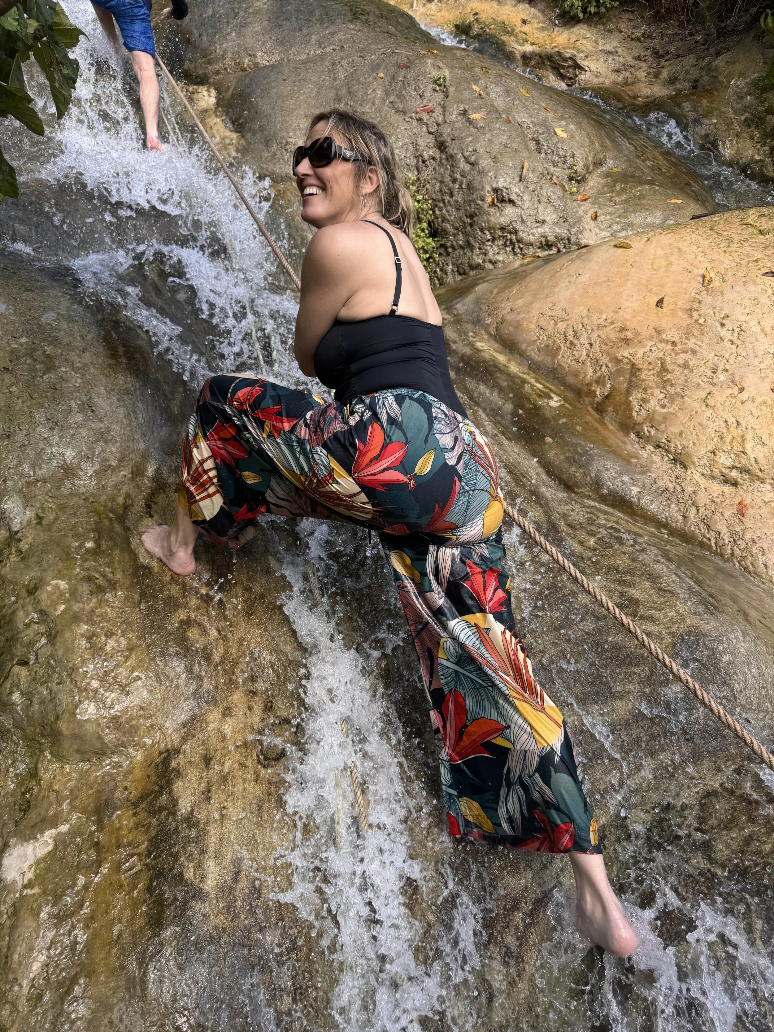 A woman smiling while climbing a rocky stream using a safety rope, wearing sunglasses, a black top, and colorful floral-patterned pants.