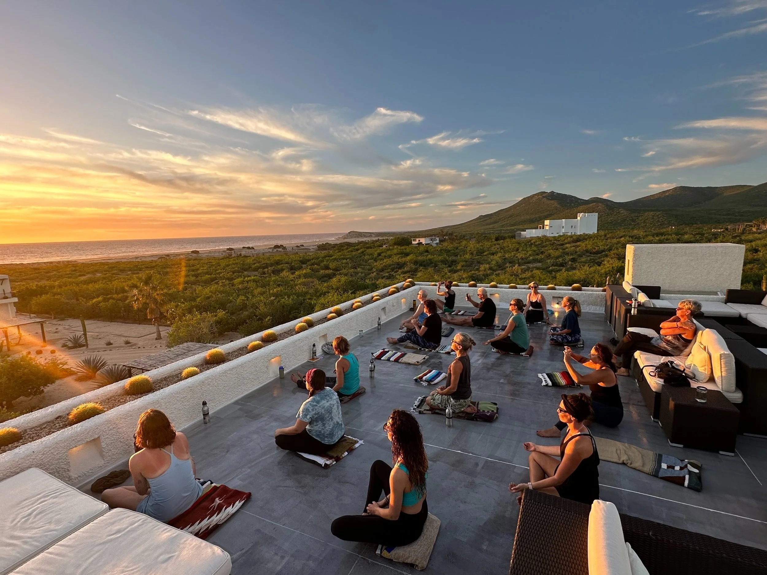 People practicing yoga on a rooftop terrace at sunset with a view of the ocean and mountains in the background.