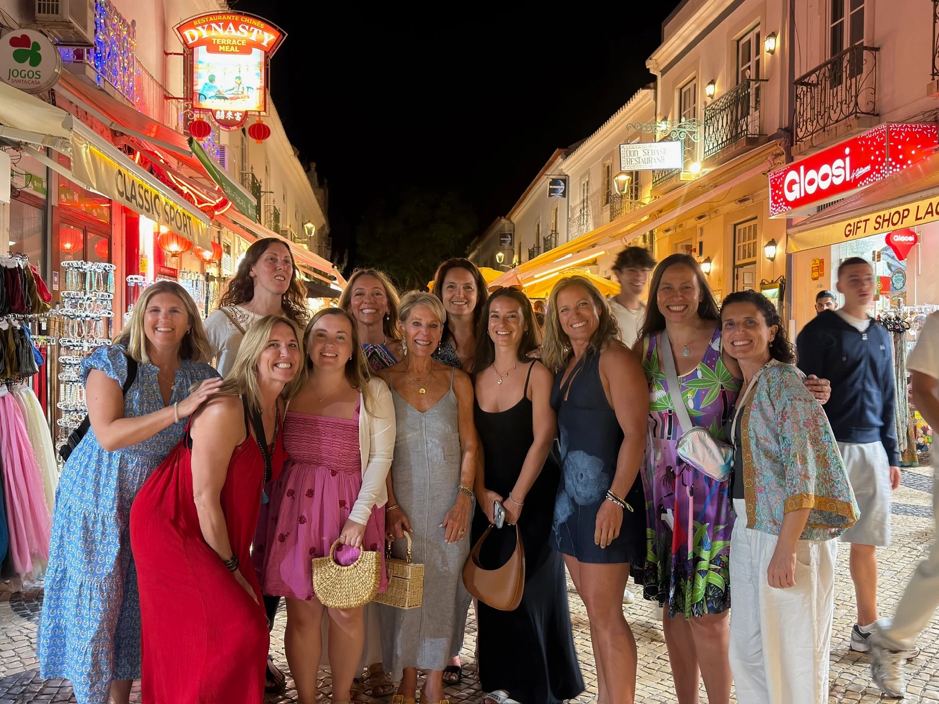 Group of women smiling and posing together on a lively city street at night, with storefronts and signage illuminated in the background.