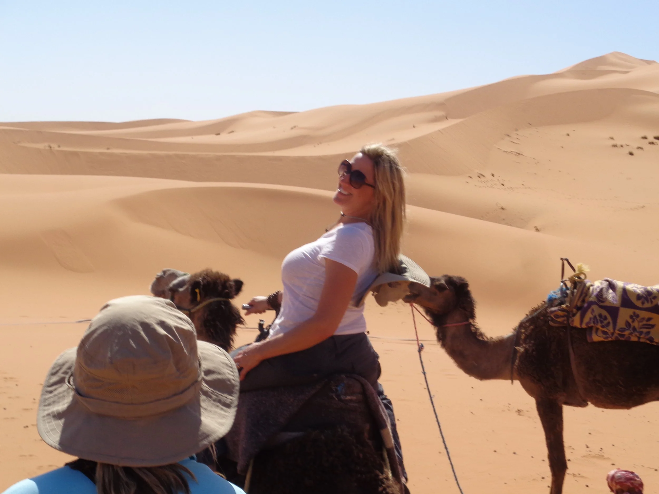 A woman smiling and wearing sunglasses, sitting on a camel in a desert with sand dunes and a clear sky, during daytime.