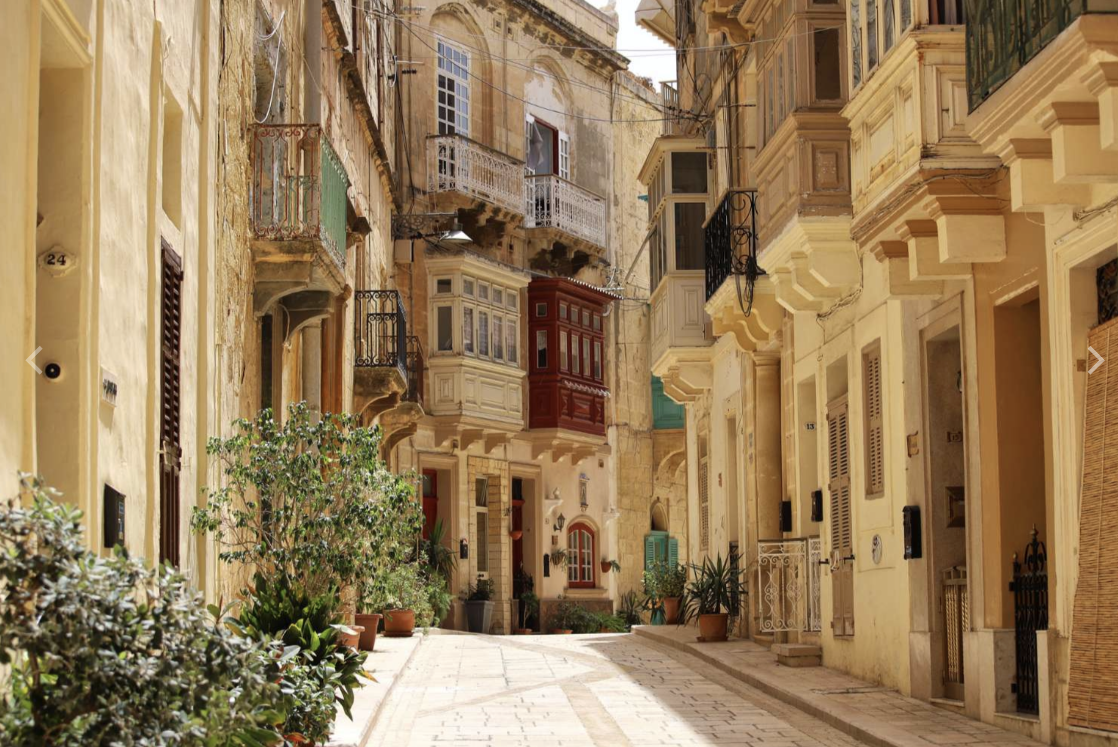 A narrow European street lined with old, yellow and beige historic buildings with balconies, windows, and outdoor plants, under a clear sky.