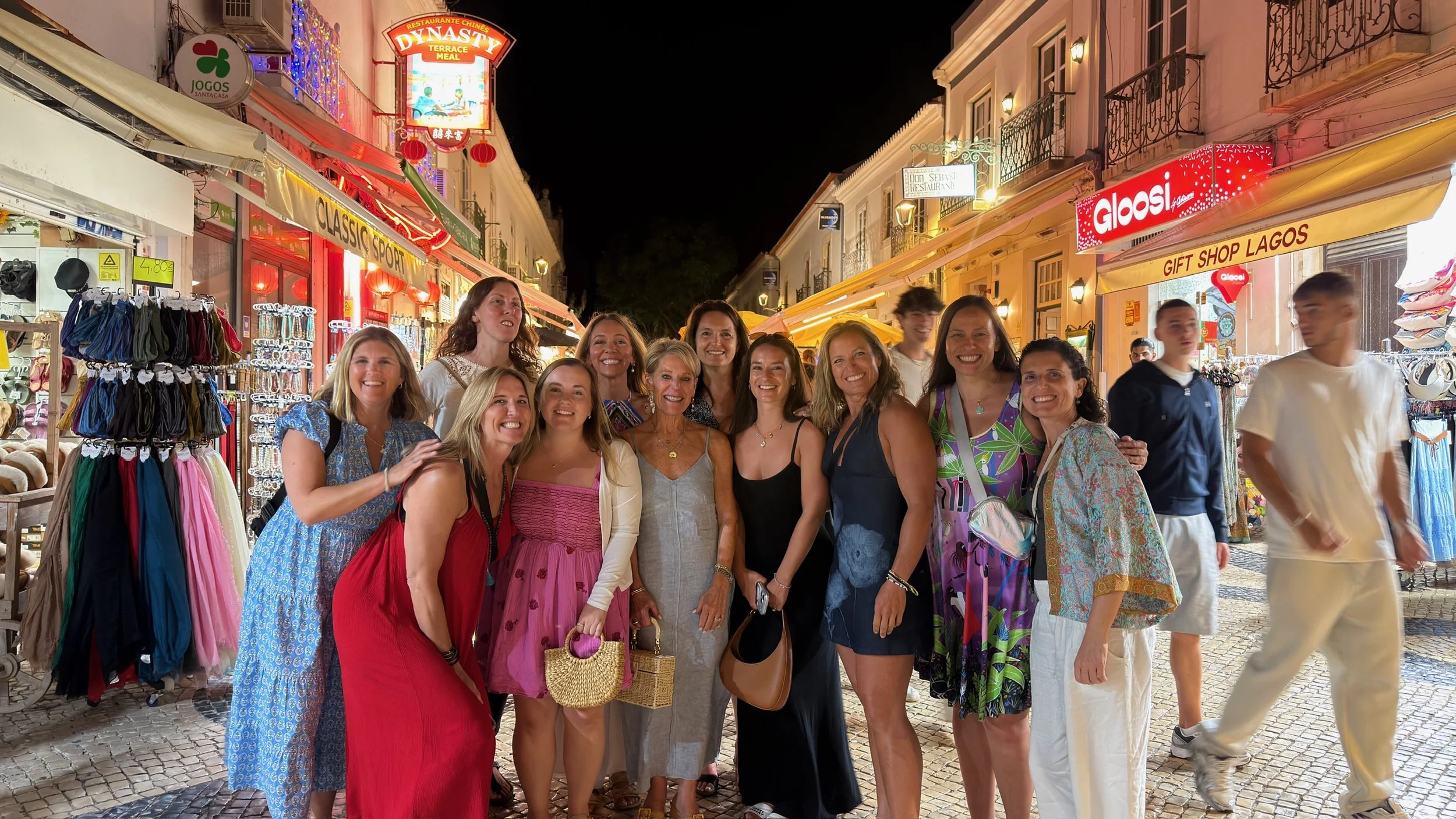 A group of women and men standing and smiling in a lively outdoor market street at night, with stores and colorful signs, including a sign that says 'Gloosi' and various shop displays.