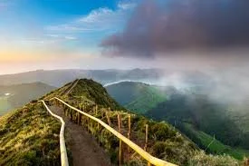 A winding mountain trail with yellow railings, lush green hills, and a cloudy sky in the distance.