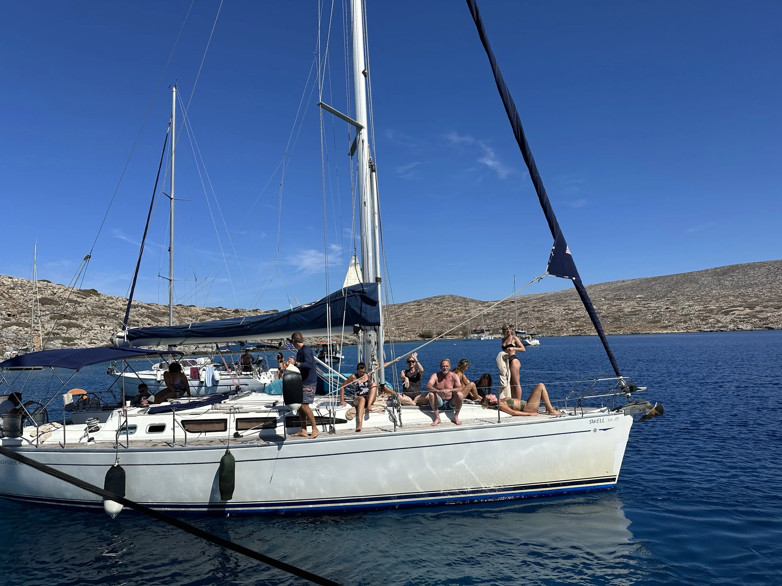 A group of people relaxing and enjoying sunny weather on a white sailboat named Swell, anchored in a calm blue body of water with hilly terrain in the background.