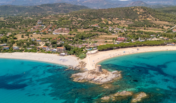 Aerial view of a beach with a stone tower on a small rocky island, turquoise water, sandy shoreline, and a coastal town with hills in the background.