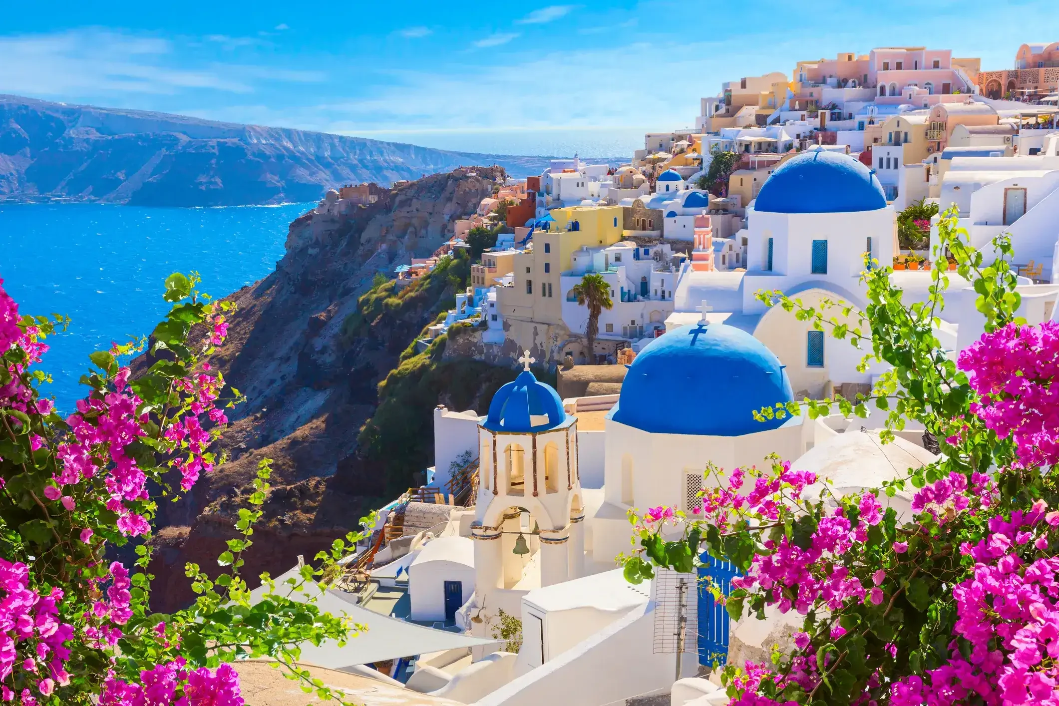 A picturesque view of Santorini, Greece, featuring white buildings with blue domes, overlooking the Aegean Sea, with pink flowers in the foreground and a cliffside landscape in the background.