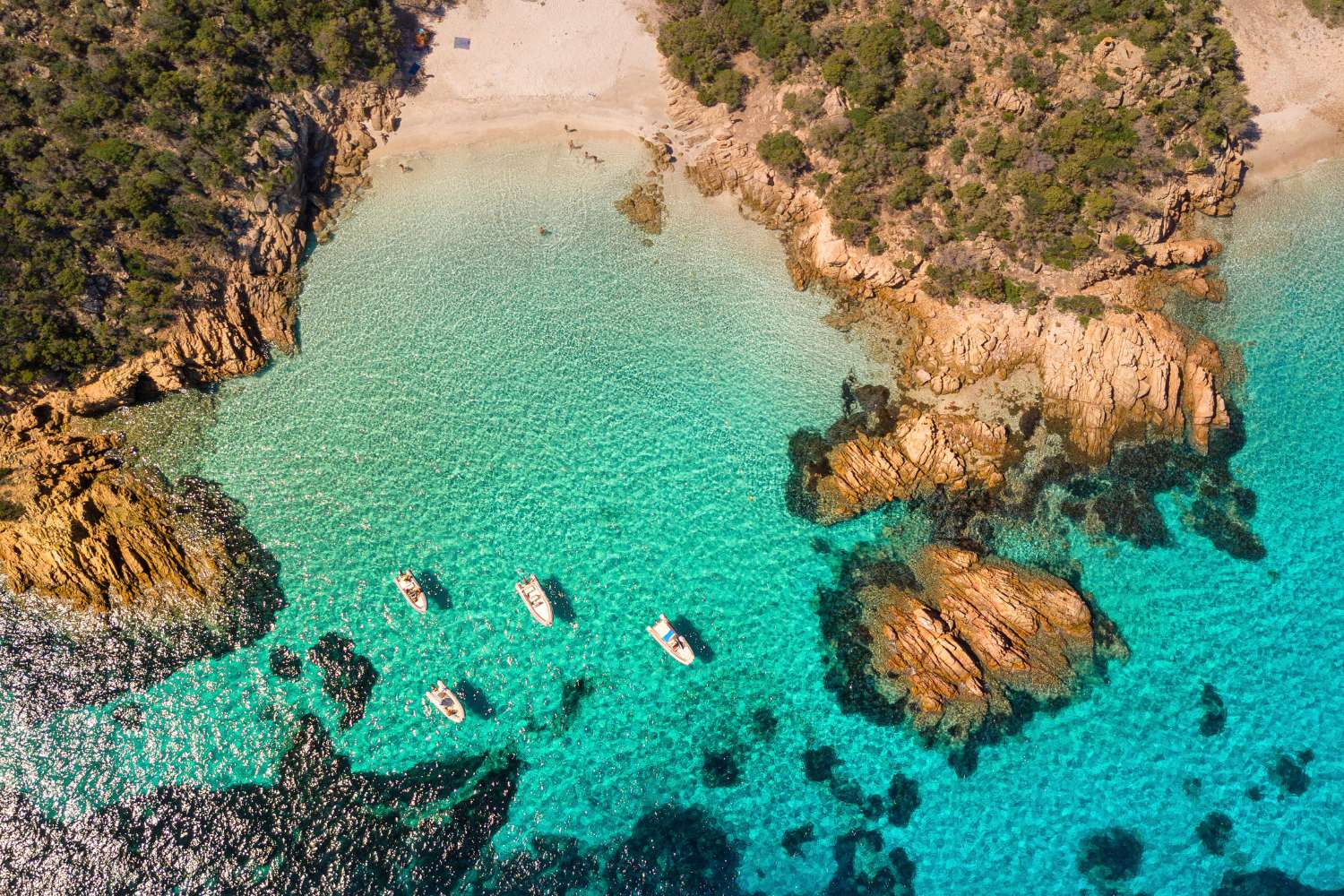 An aerial view of a secluded cove with turquoise water, rocky cliffs, a sandy beach, and several boats anchored in the water.