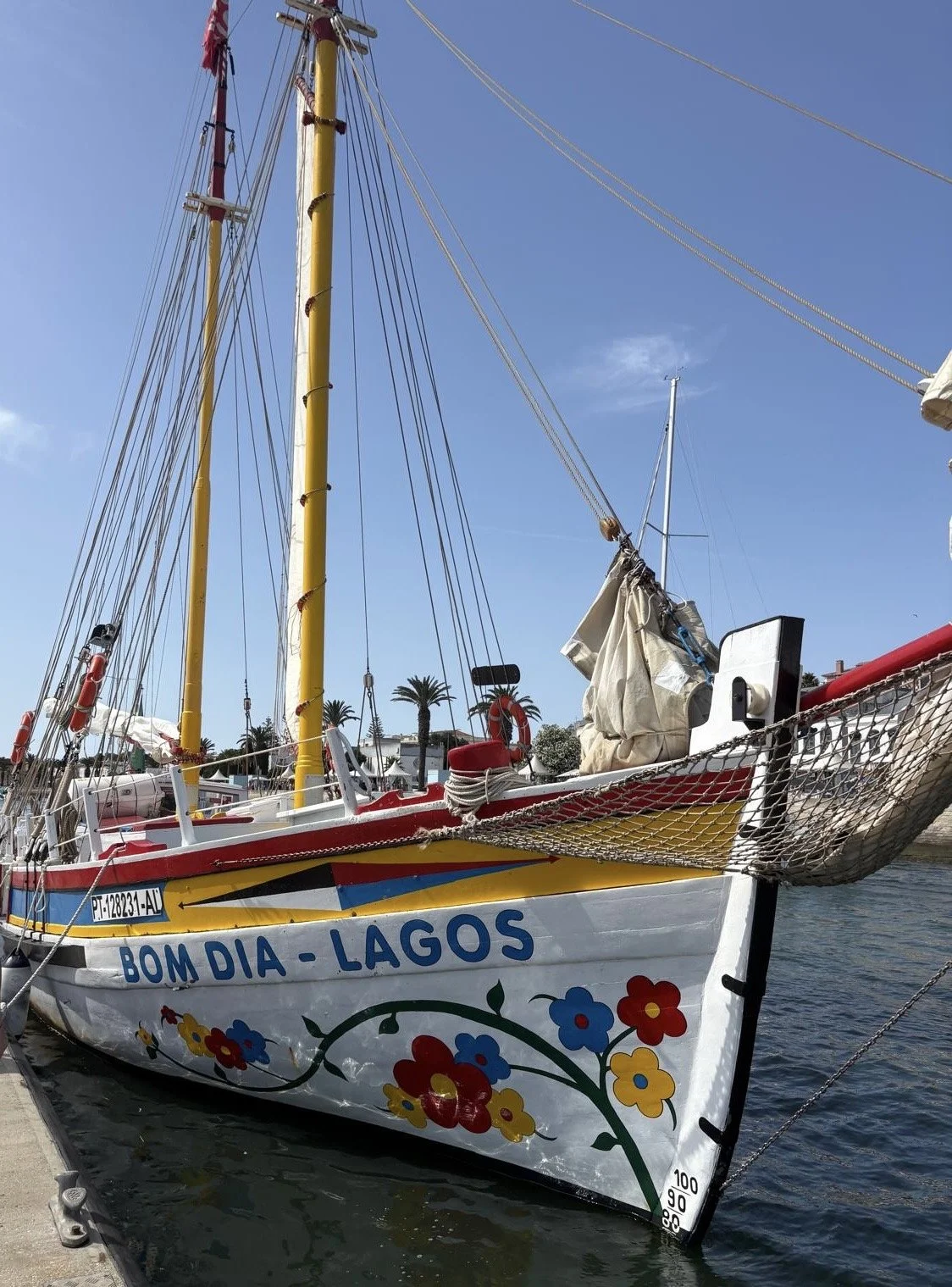 Colorful sailboat named 'Bom Dia - Lagos' docked at marina, with bright blue sky and palm trees in background, decorated with painted flowers on the hull.